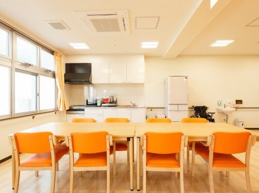 オレンジの椅子がセットされたダイニングテーブルが手前にあり、奥には冷蔵庫や本格的なキッチンがある。