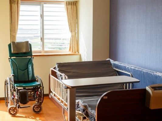 ネイビーブルーのアクセントクロスが貼られたフローリングの部屋には介護ベッドや車いすが置かれている。