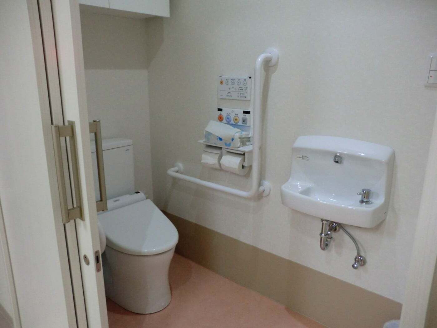 手洗い場や手すり、操作ボタンが壁に付いたトイレは広く、出入り口には引き戸が設置されている。