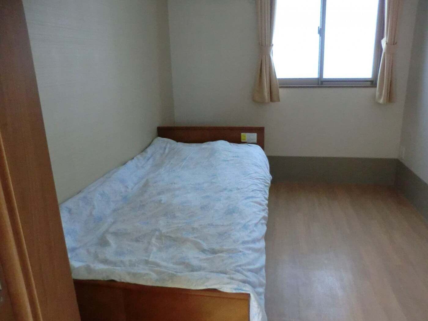 フローリングの部屋はコンパクトなサイズで、寝具付きのベッドが置かれ、窓にはカーテンがある。
