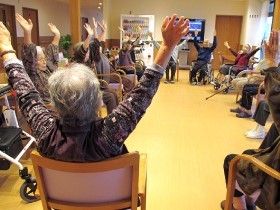 体操をしている様子の高齢者。施設内の広々とした空間に椅子を並べて集まっている