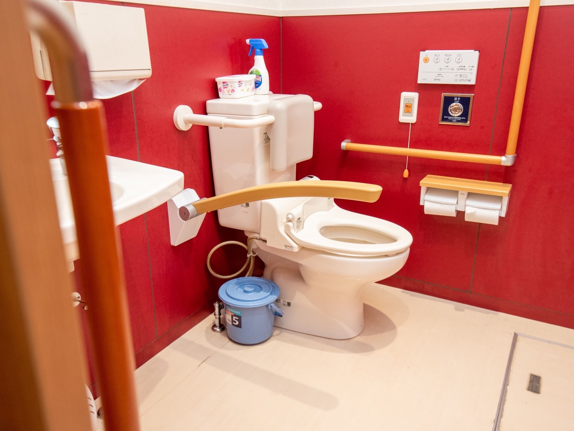 赤い壁に囲まれた広いトイレがある。壁には手すりが設置され、可動式のひじ掛けが付いている。側には洗面台とペーパーホルダーがある。
