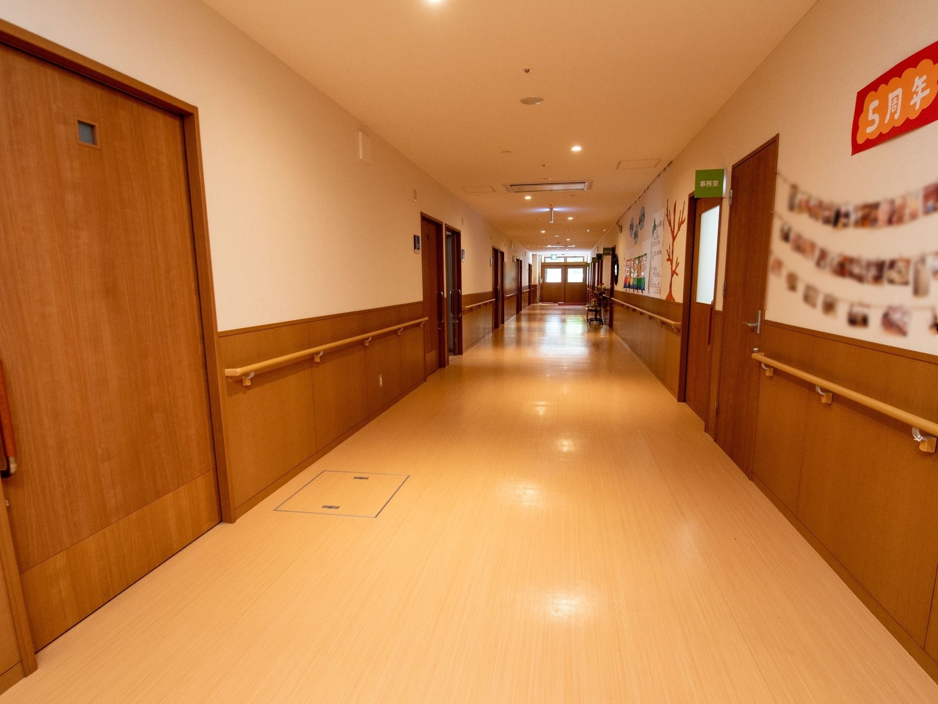 幅の広いベージュの廊下がある。両隣には木目調の居室のドアが並んでいる。壁には手すりがあり、写真や飾りつけがされている。