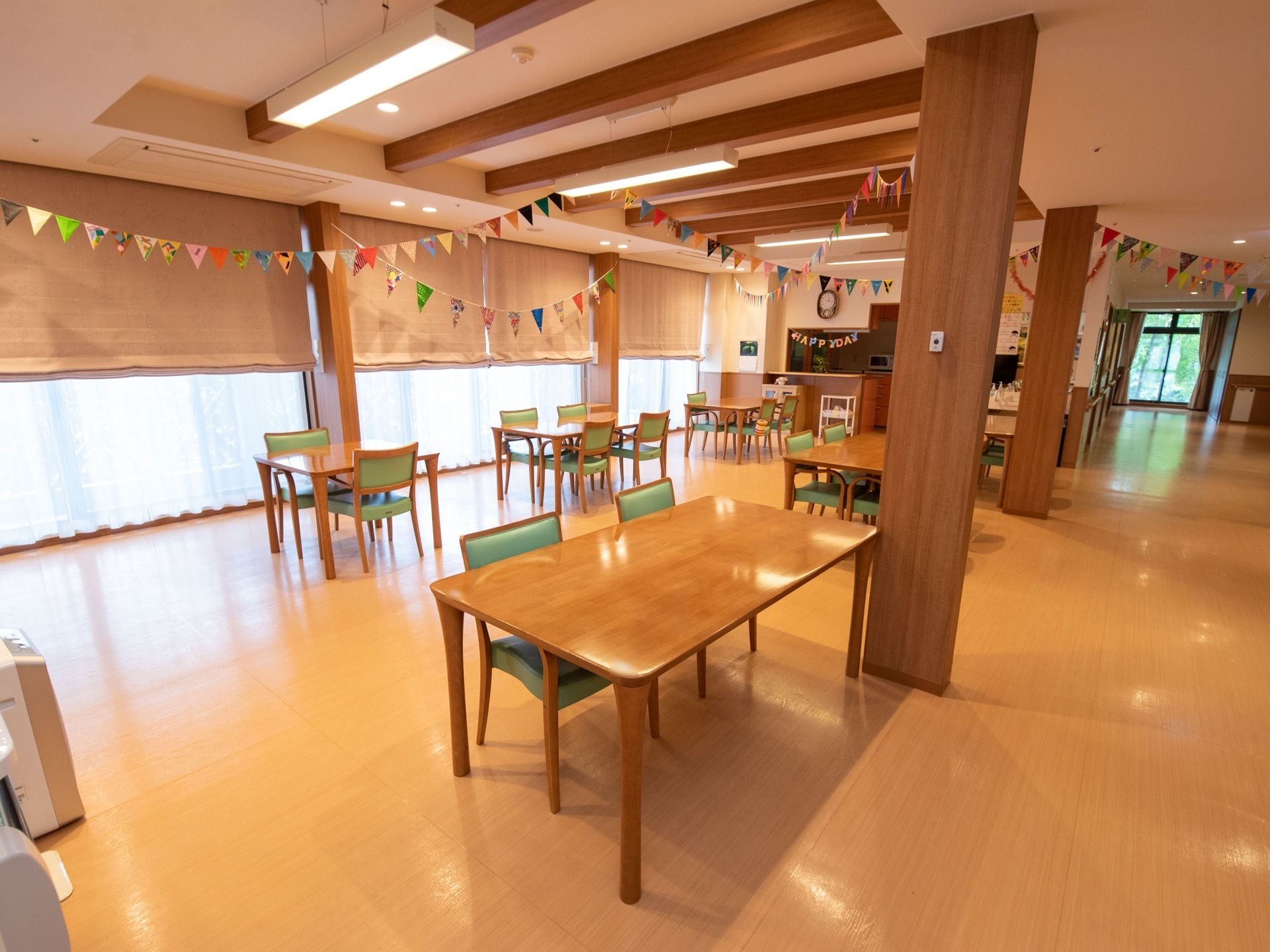 木の柱と梁が渡されている広い食堂がある。壁一面は窓になっており、ブラインドが下がっている。テーブルと椅子がたくさん置かれている。