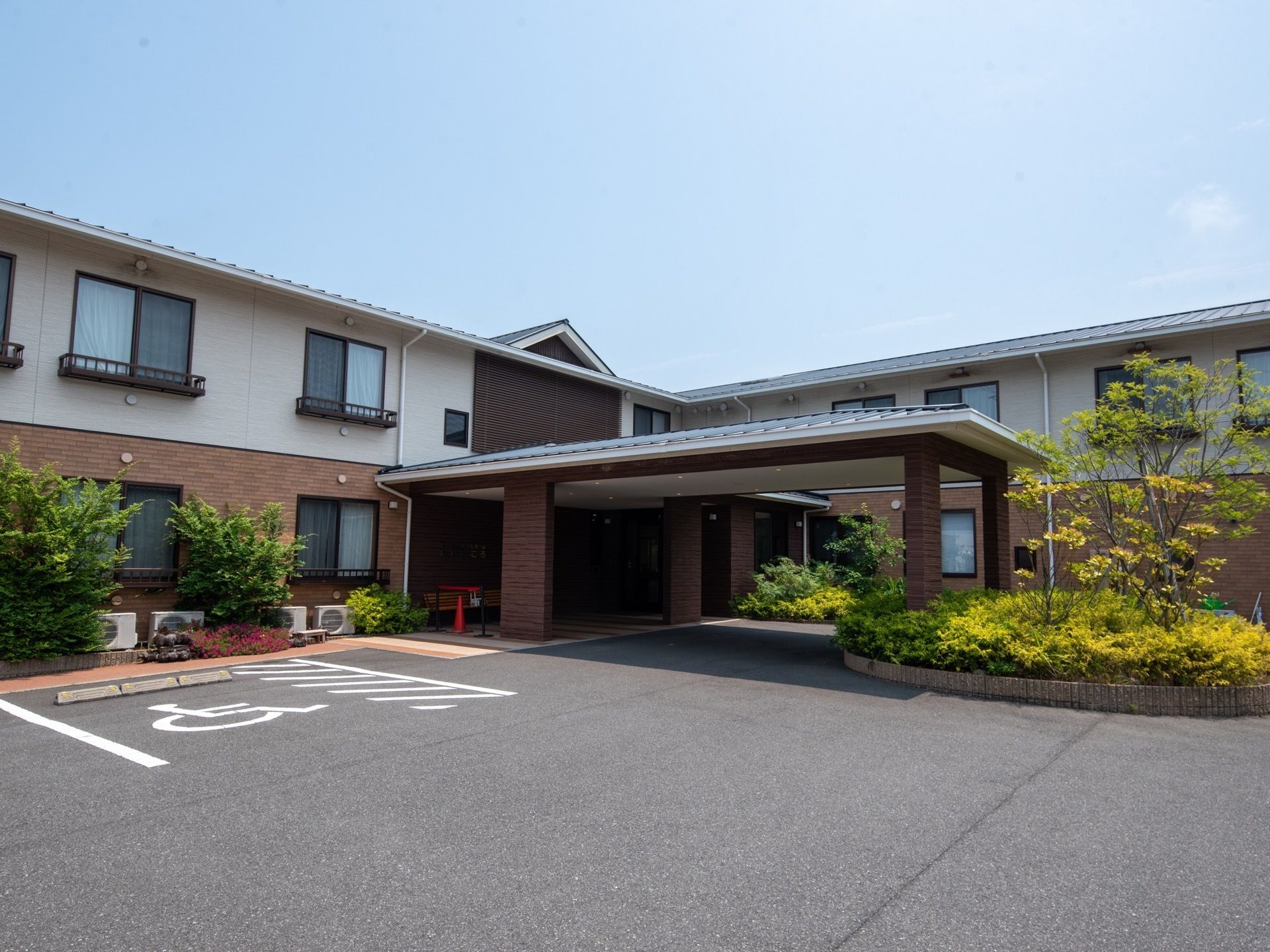 ２階建てのL字型の建物がある。角の部分にルーフが設置された玄関がある。周囲には木がたくさん植えられており、周辺は駐車場になっている。