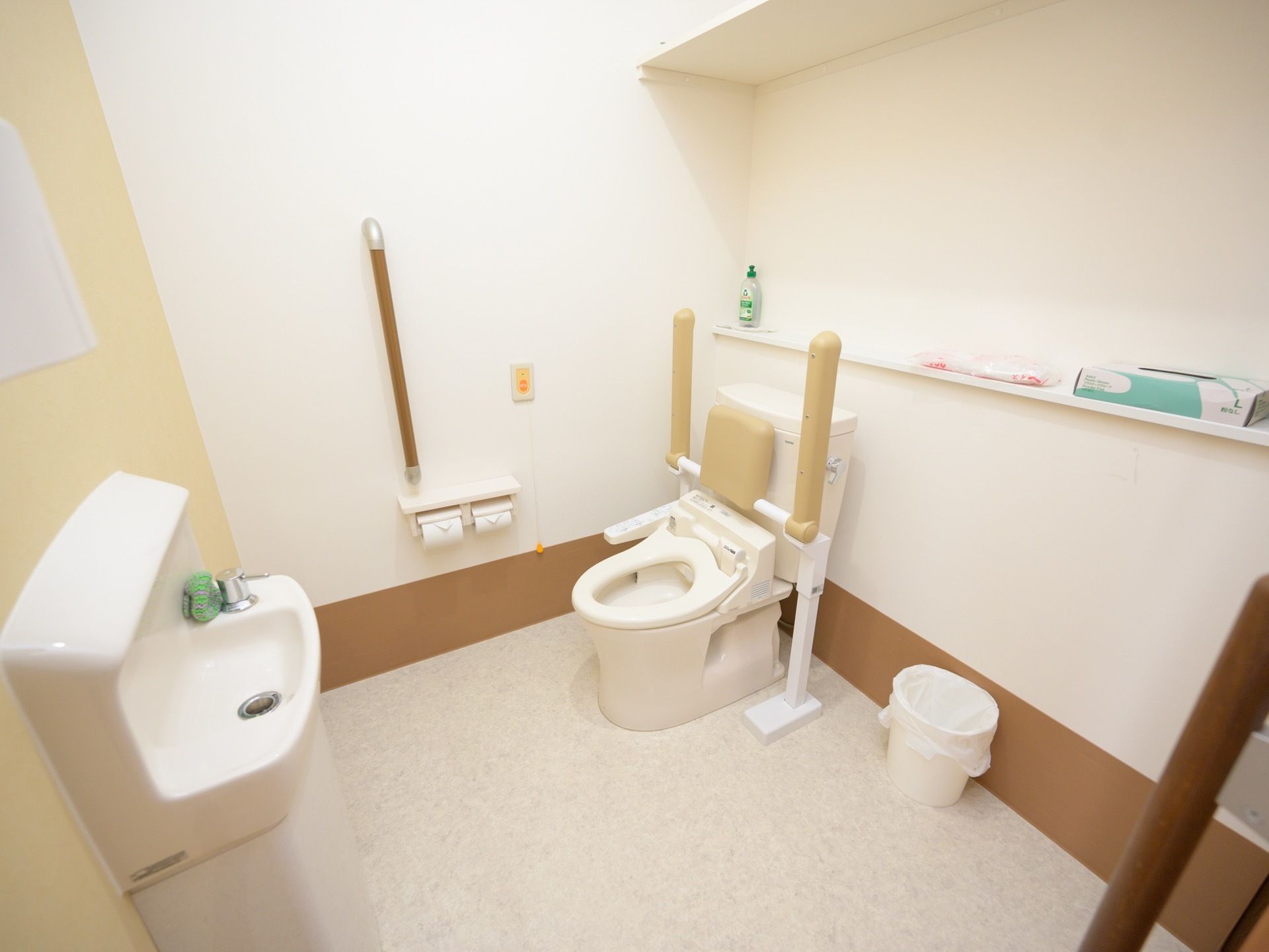 トイレには洋式便器があり、前方は広く空いている。便器には可動式手すり、壁には固定された手すりがある。呼び出しボタンもある。