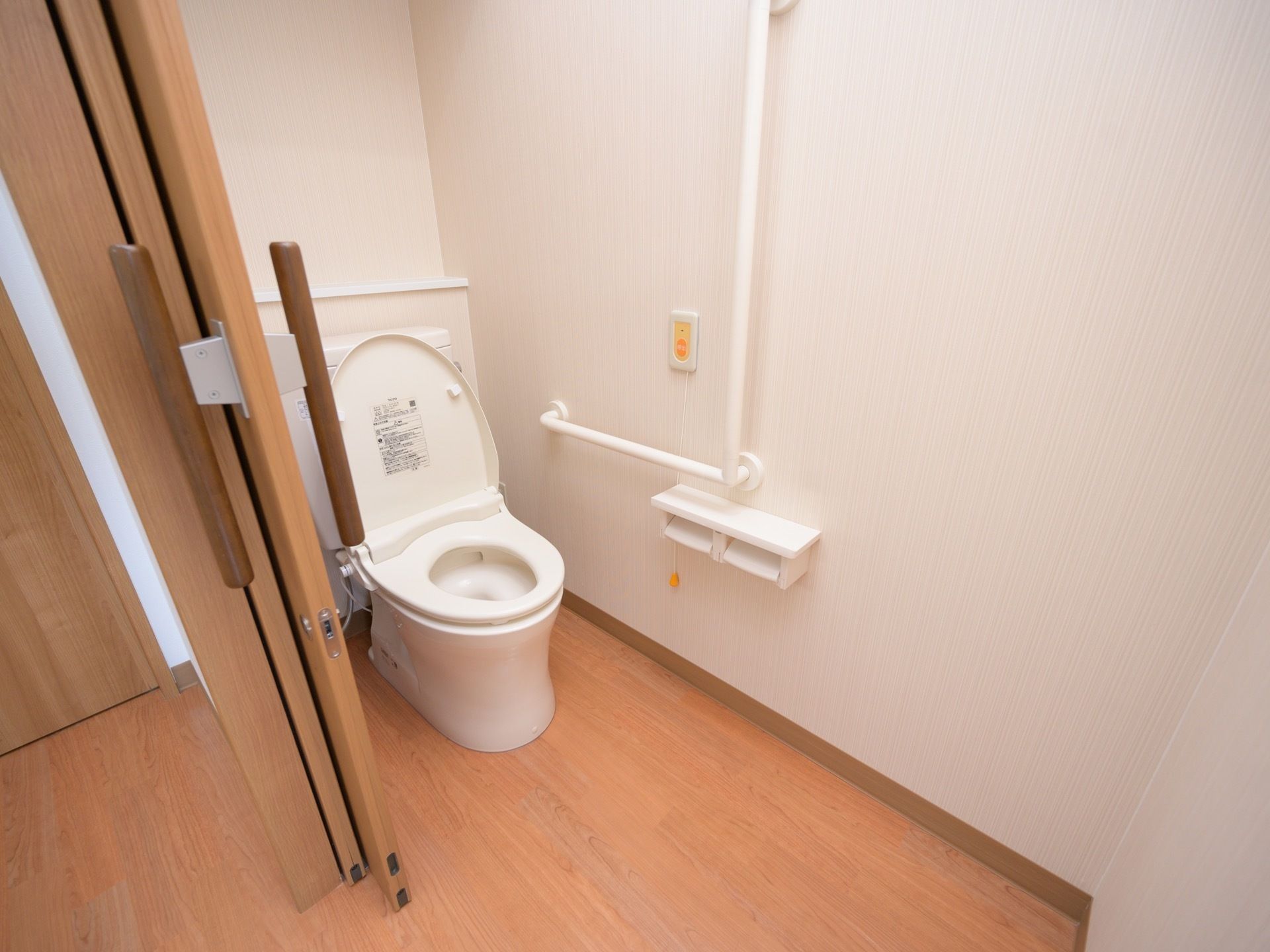 トイレには洋式便器があり、壁にはトイレットペーパーフォルダーと長い手すり、呼び出しボタンがある。