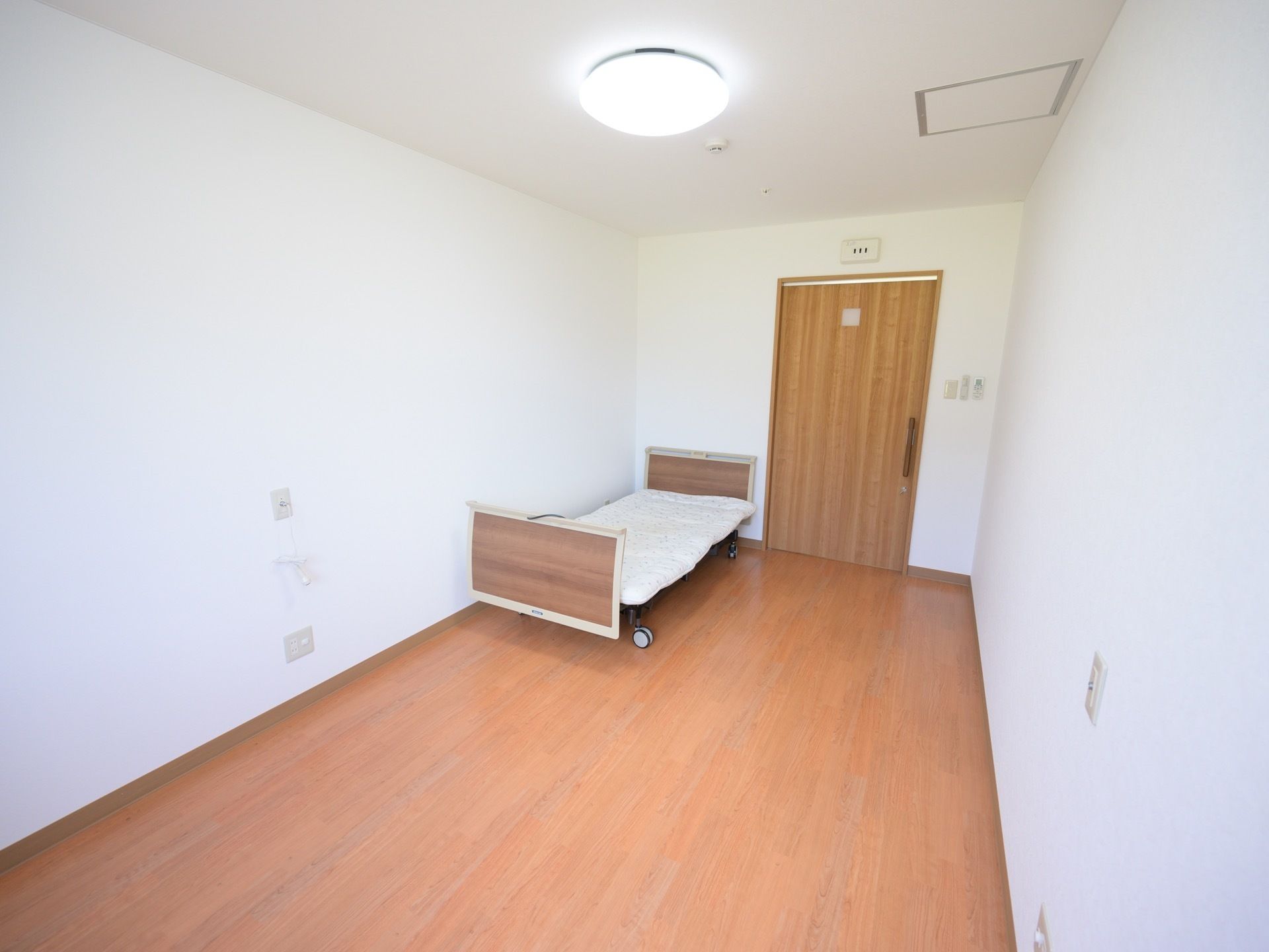 居室は白い壁で、フローリングの床となっている。茶色い扉の横側に、ベッドを用意。照明が点灯している。