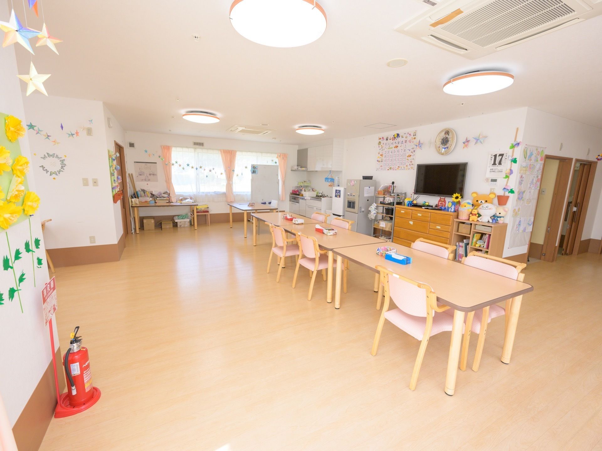 部屋の中央に３台のテーブル、複数の椅子が並べられている。キッチンスペースもあり、冷蔵庫が置かれている。