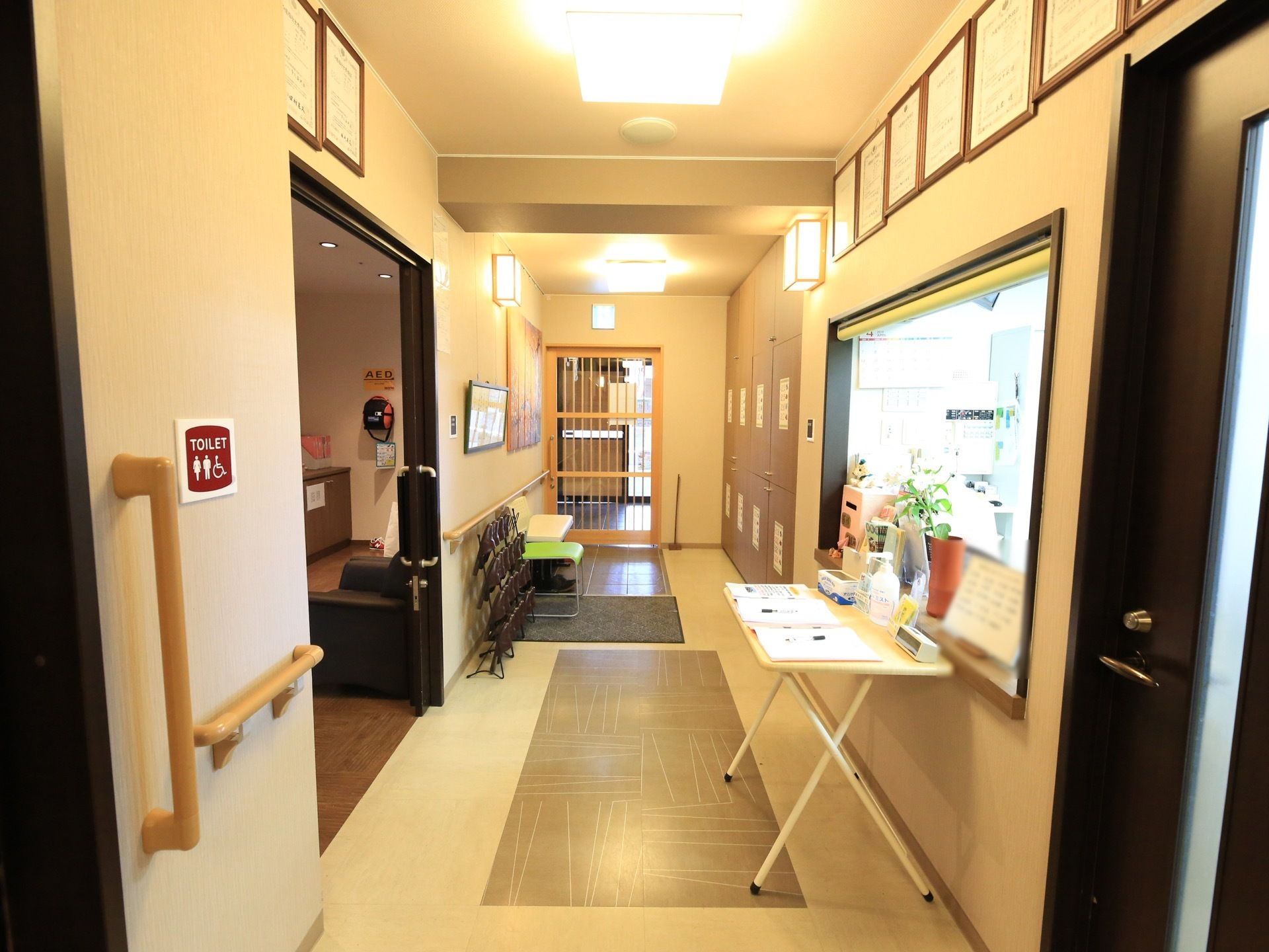 出入り口の近くに受付窓口があり、観葉植物などが置かれている。前に机が置かれていて、書類や消毒用のボトルが乗っている。