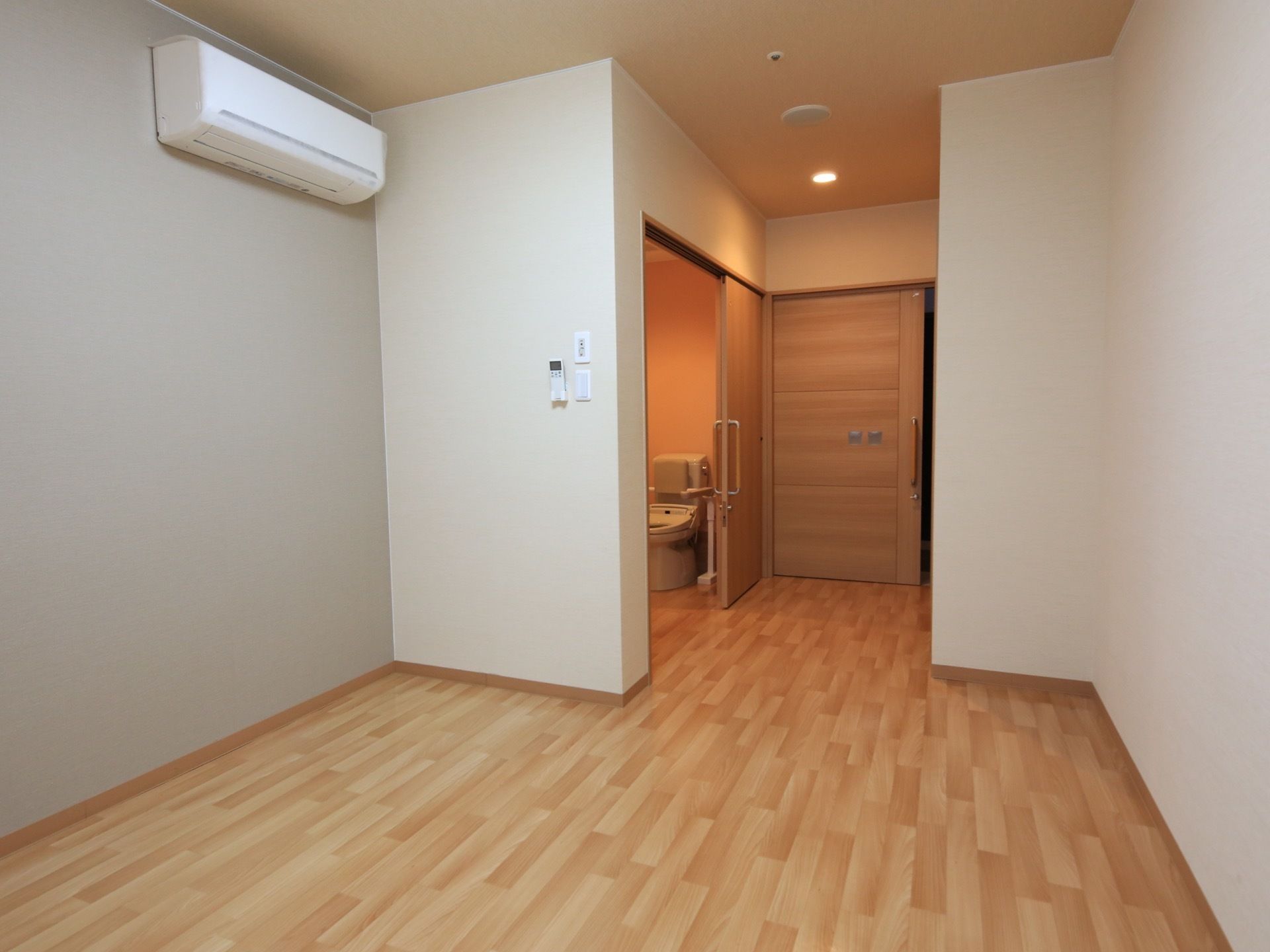 居室内にあるトイレのドアが開いていて、床面はバリアフリーとなっている。壁には白いエアコンが取りつけられている。