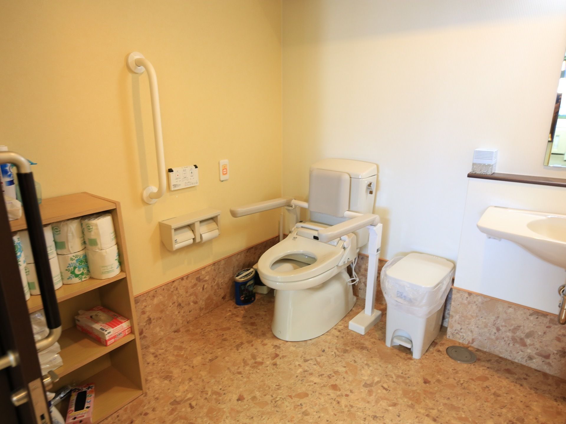 トイレには肘掛けと背もたれが取りつけられている。手前に棚があり、トイレットペーパーなどが入っている。