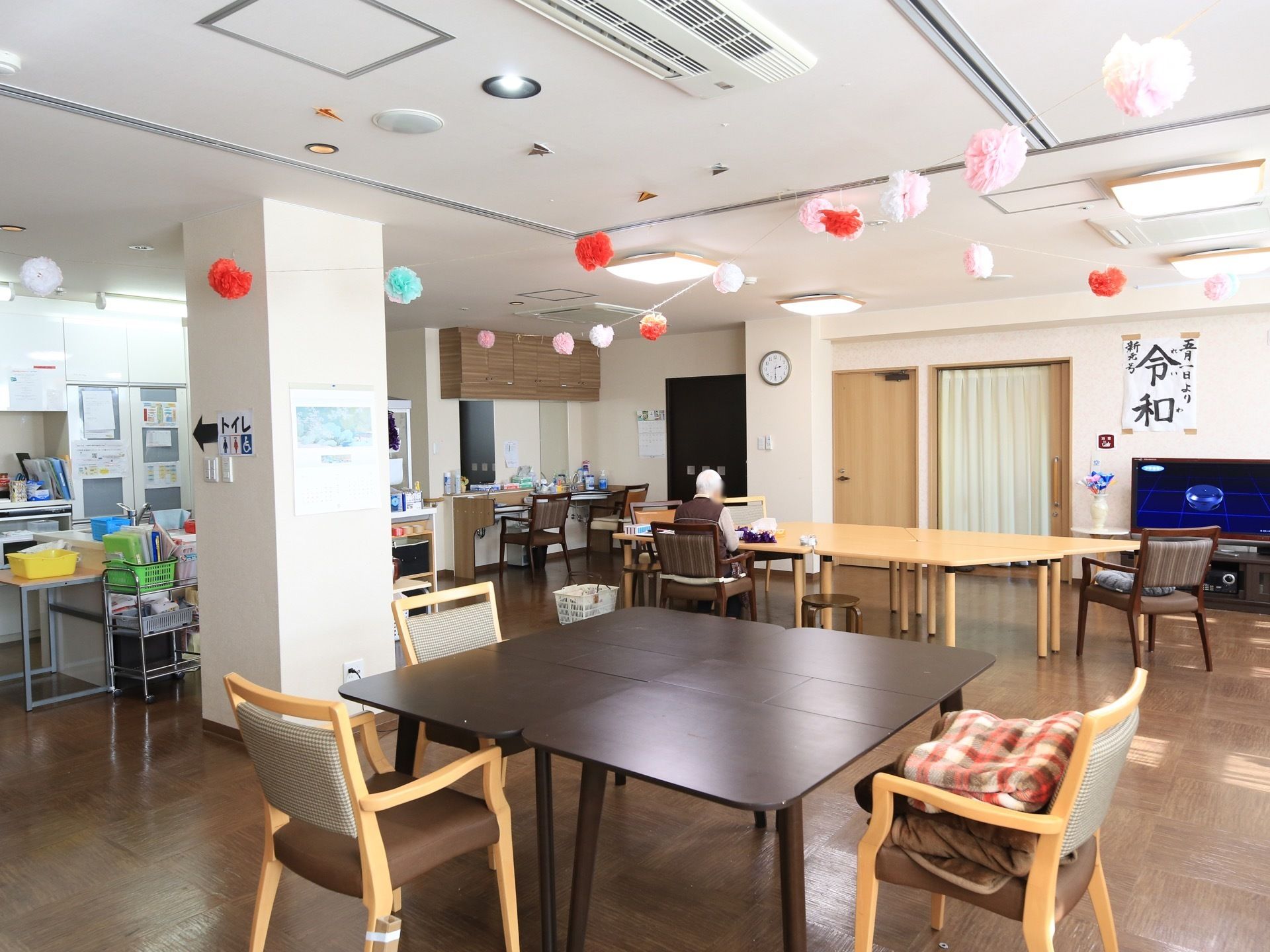 食堂で奥のテーブルに一人の入居者様が座っているのが見える。天井にはカラフルな花の飾りがつけられている。