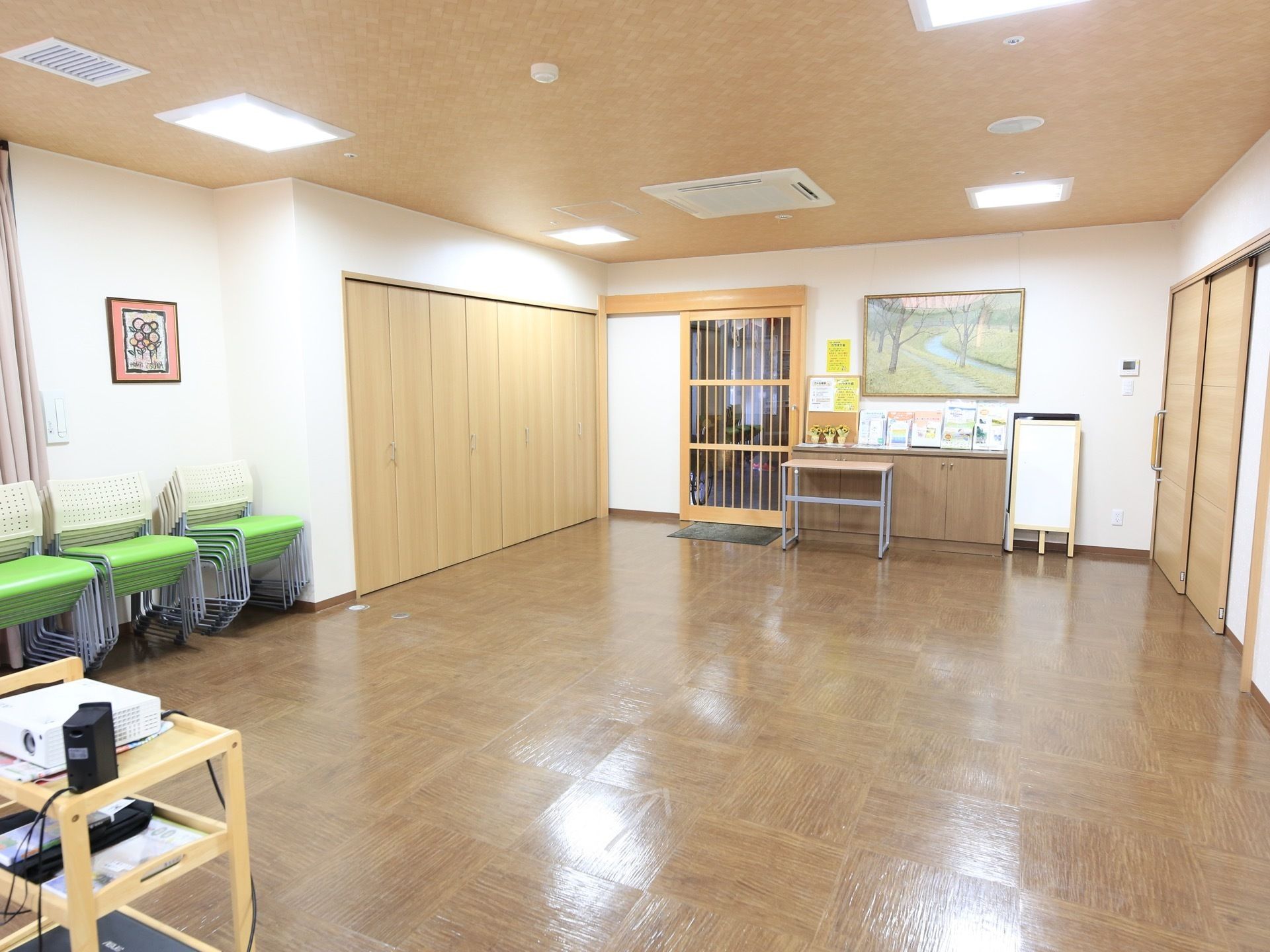 出入り口に木の格子戸が設置されていて、壁には絵画が飾られている。壁際にグリーンの椅子が多数重ねて置かれている。
