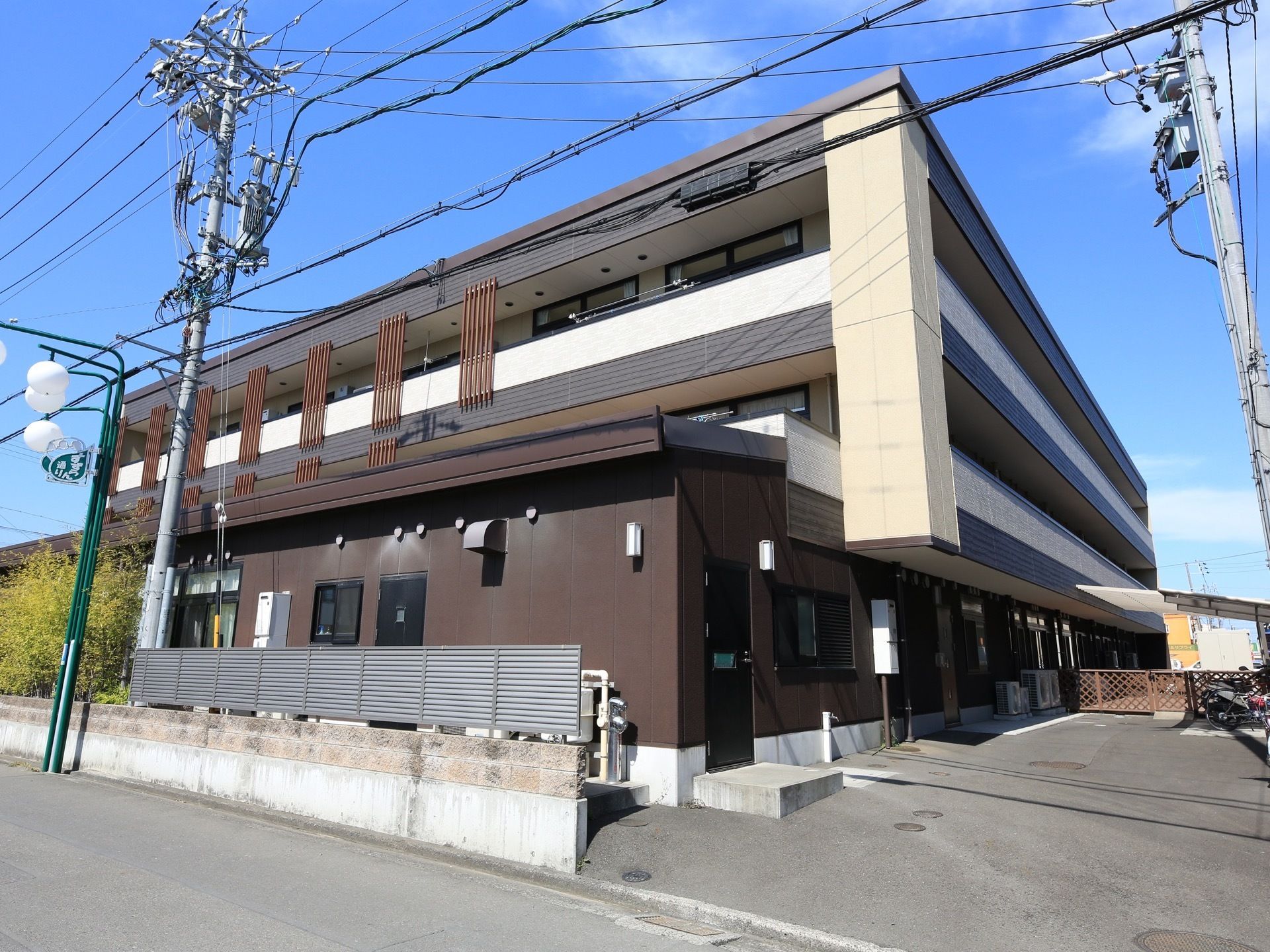 ３階建ての建物で外壁は濃いブラウンである。建物の前にはグレーの柵があり、外側に緑色の街灯が立っている。