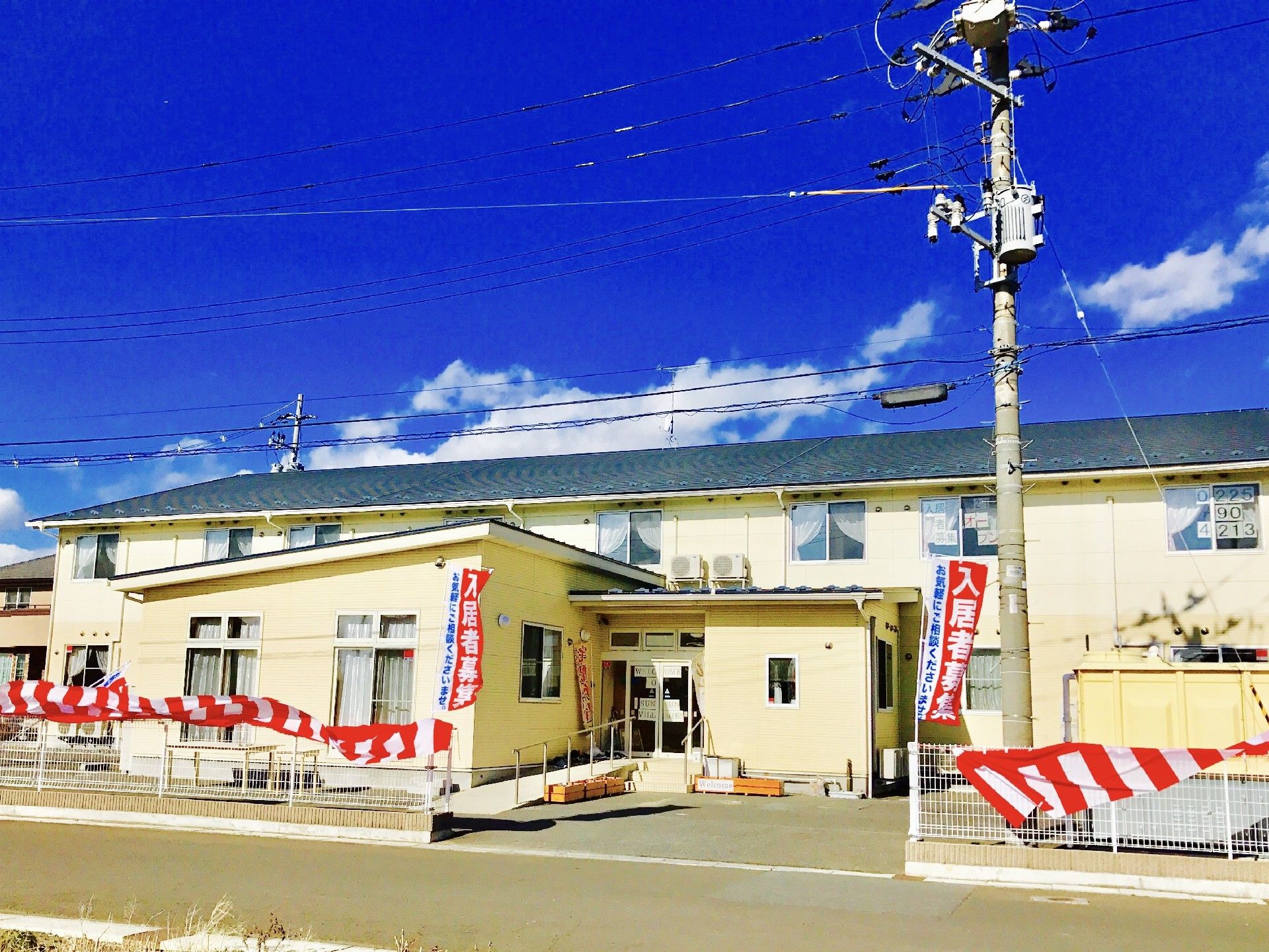 紅白の垂れ幕が建物周辺のフェンスにかかった2階建ての建物。その前に平屋建てもある。