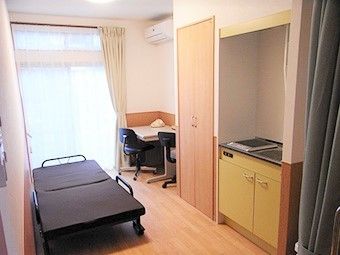 居室のベッドの右奥にはテーブルがあり、手前にはキッチン、奥の窓にはカーテンが付けられている。