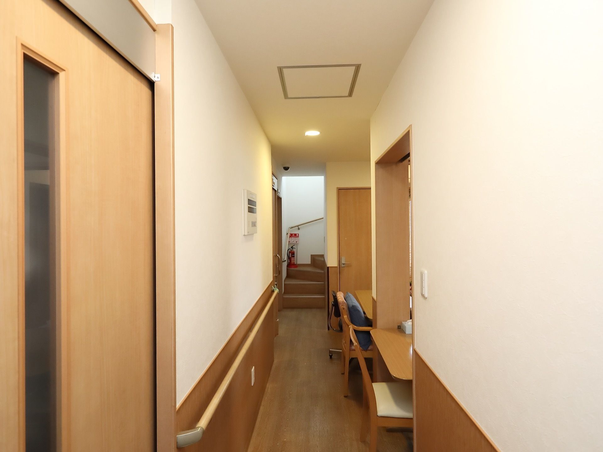ナチュラル系の木材の腰壁が設置された通路は、あたたかみがある。壁に沿って手すりが取りつけられている。階段に消火設備が置かれている。