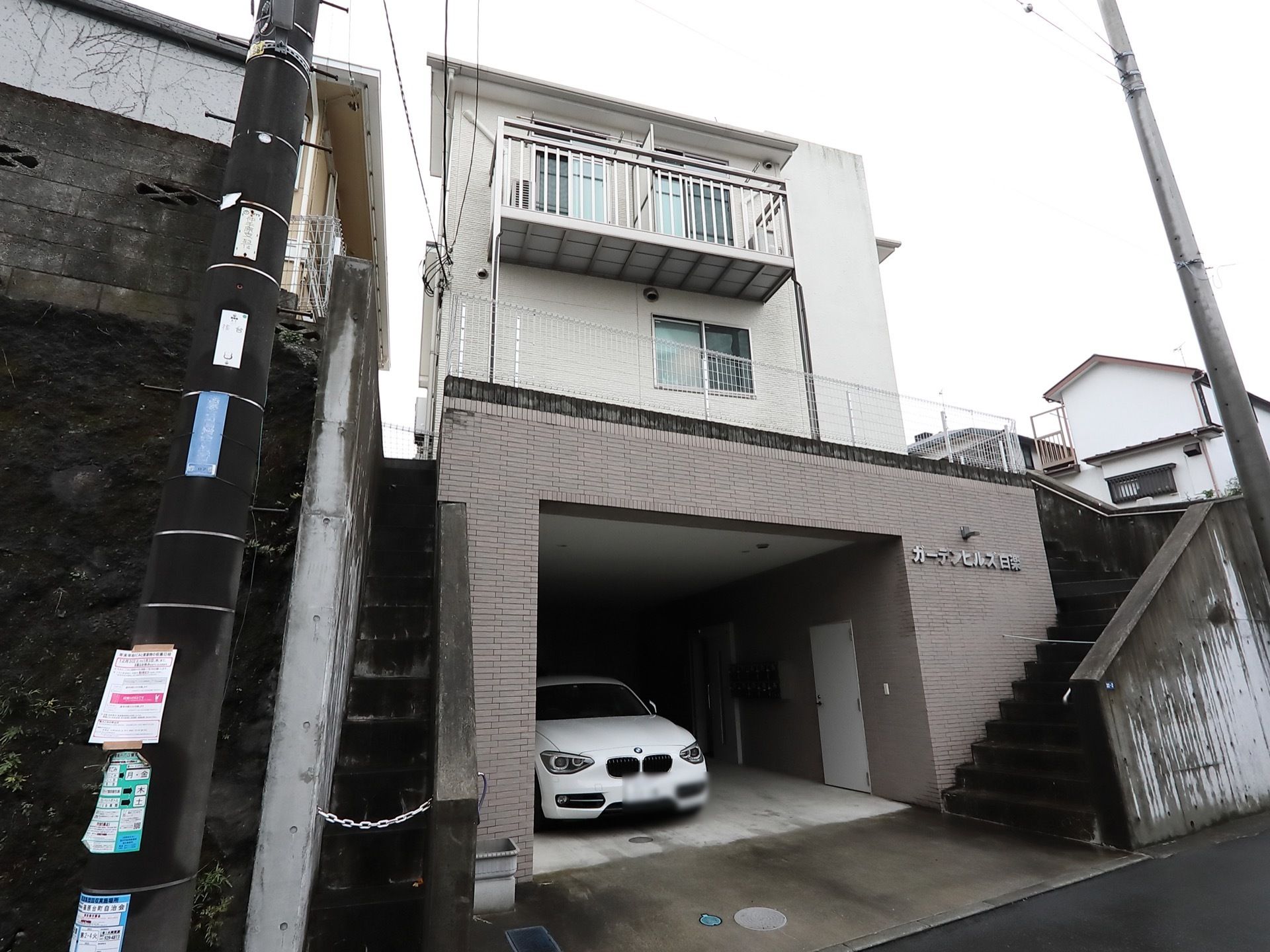 施設は１階部分が駐車スペースになっている。建物に向かって右側に階段がある。建物の外壁は白を基調としている。