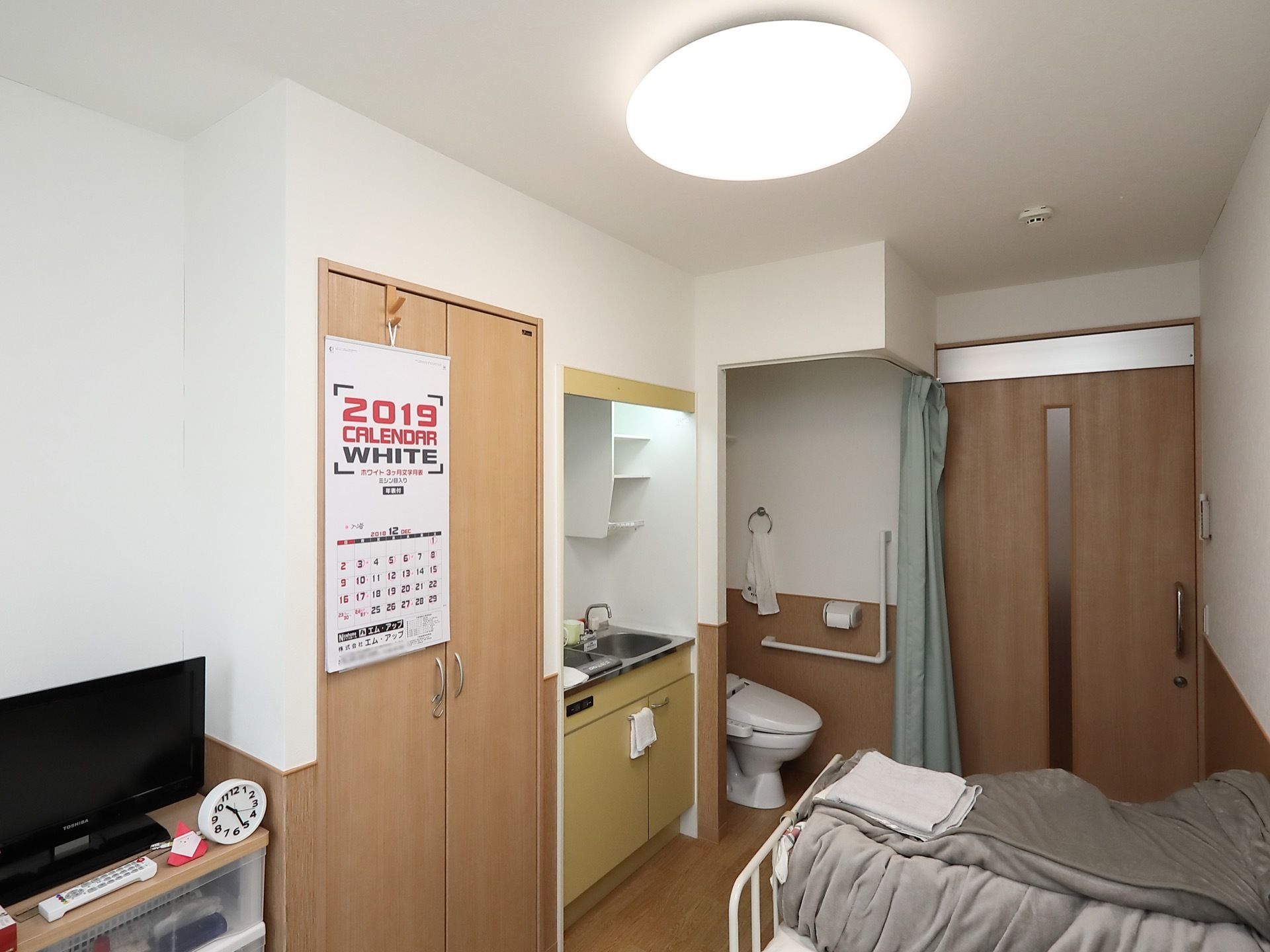 洋室の居室で、ベッドや収納棚などの家具やテレビが置かれている。キッチンやクローゼットは備えつけである。