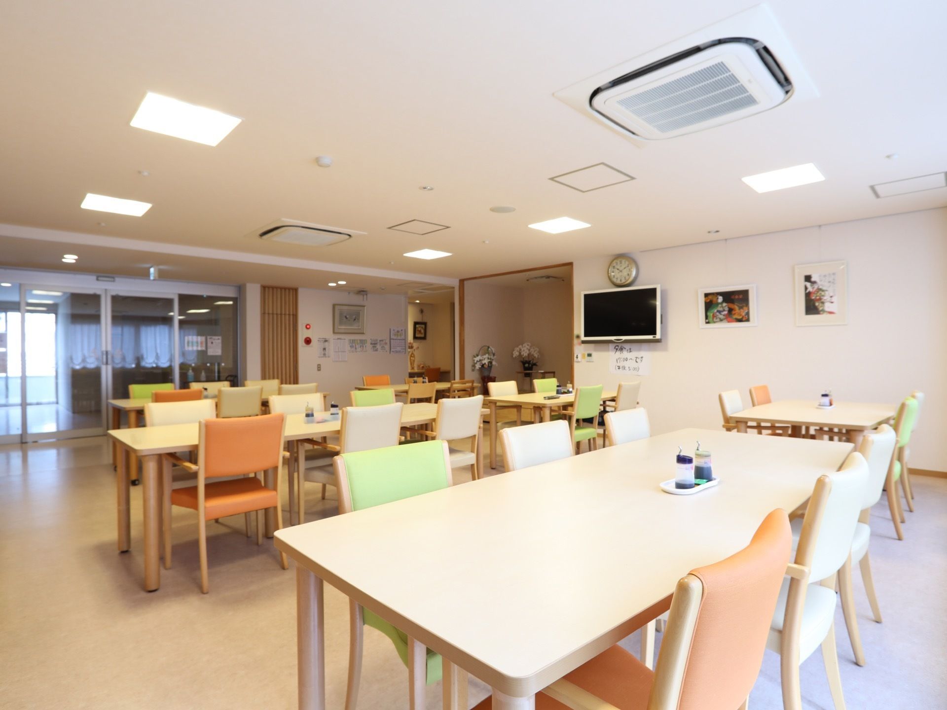オレンジとグリーンとホワイトの椅子が並んだ食堂。テーブルの上には調味料が置かれており、壁にはテレビが備え付けられている。