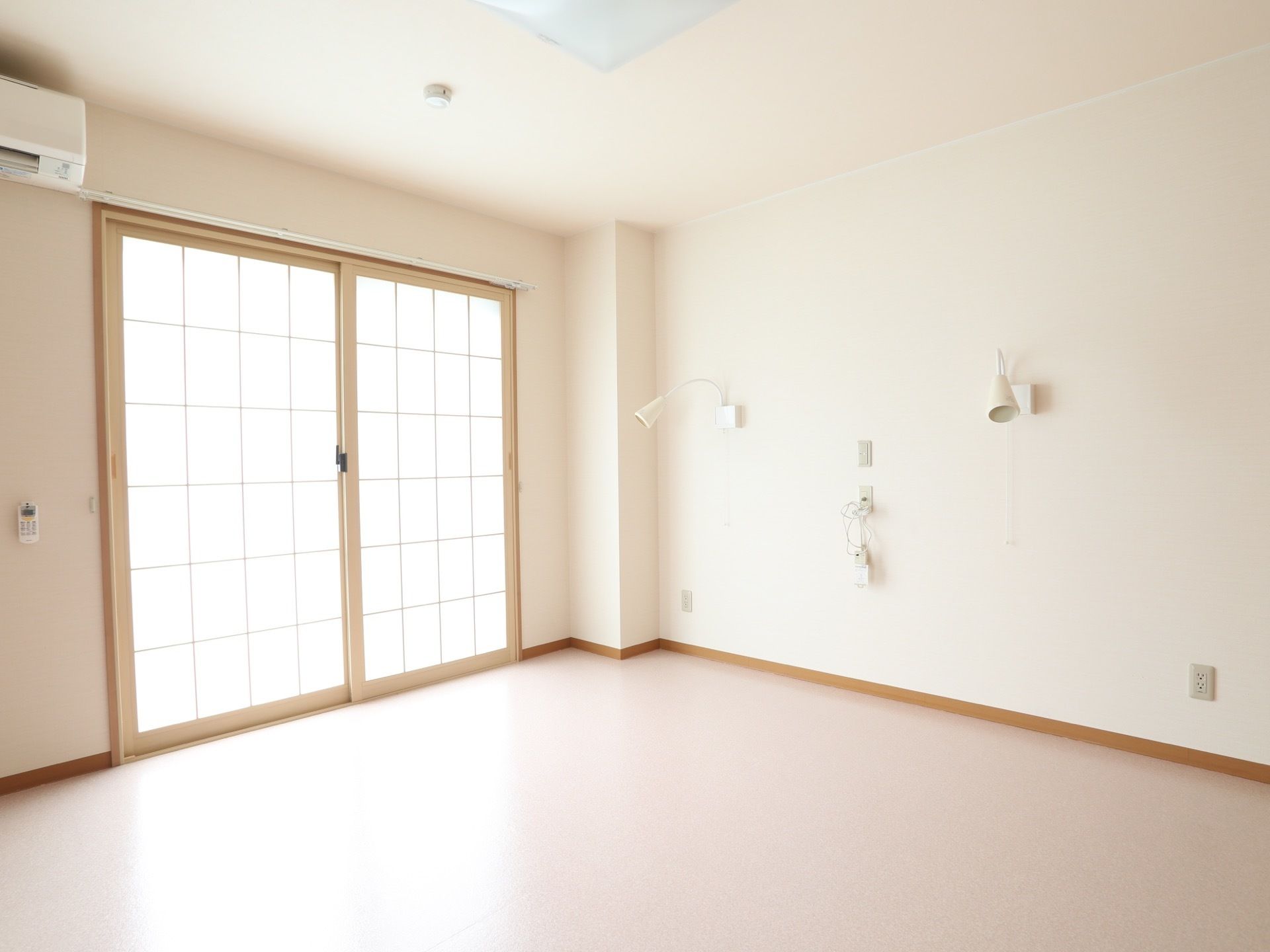 障子風の掃き出し窓が付いた明るい居室。窓の上にはエアコンが設置されており、壁には照明とナースコールが付いている。