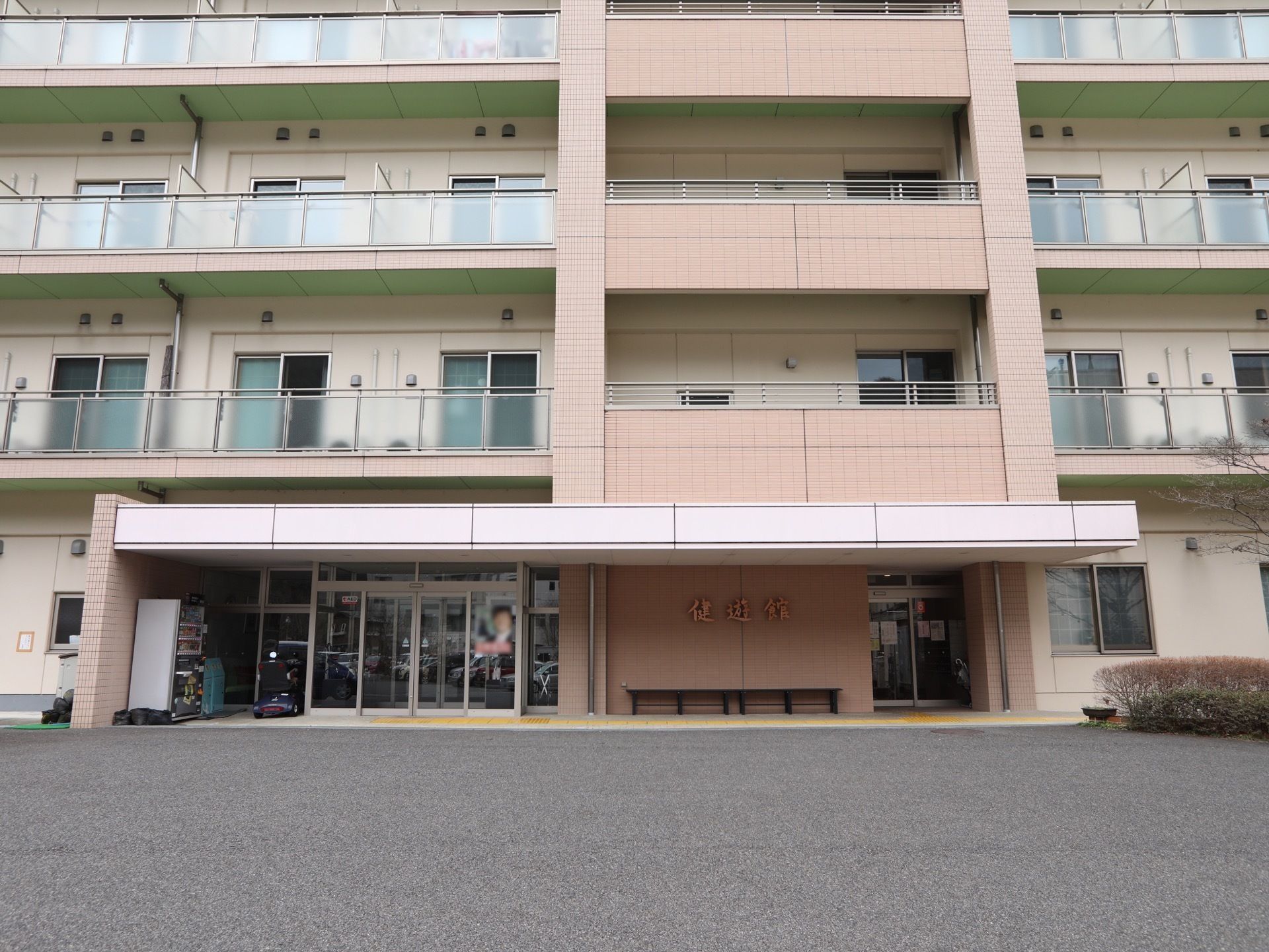 アスファルトで舗装された敷地に施設の建物が建っている。入口は両開きの自動ドアで、ドアの横には自動販売機が設置されている。