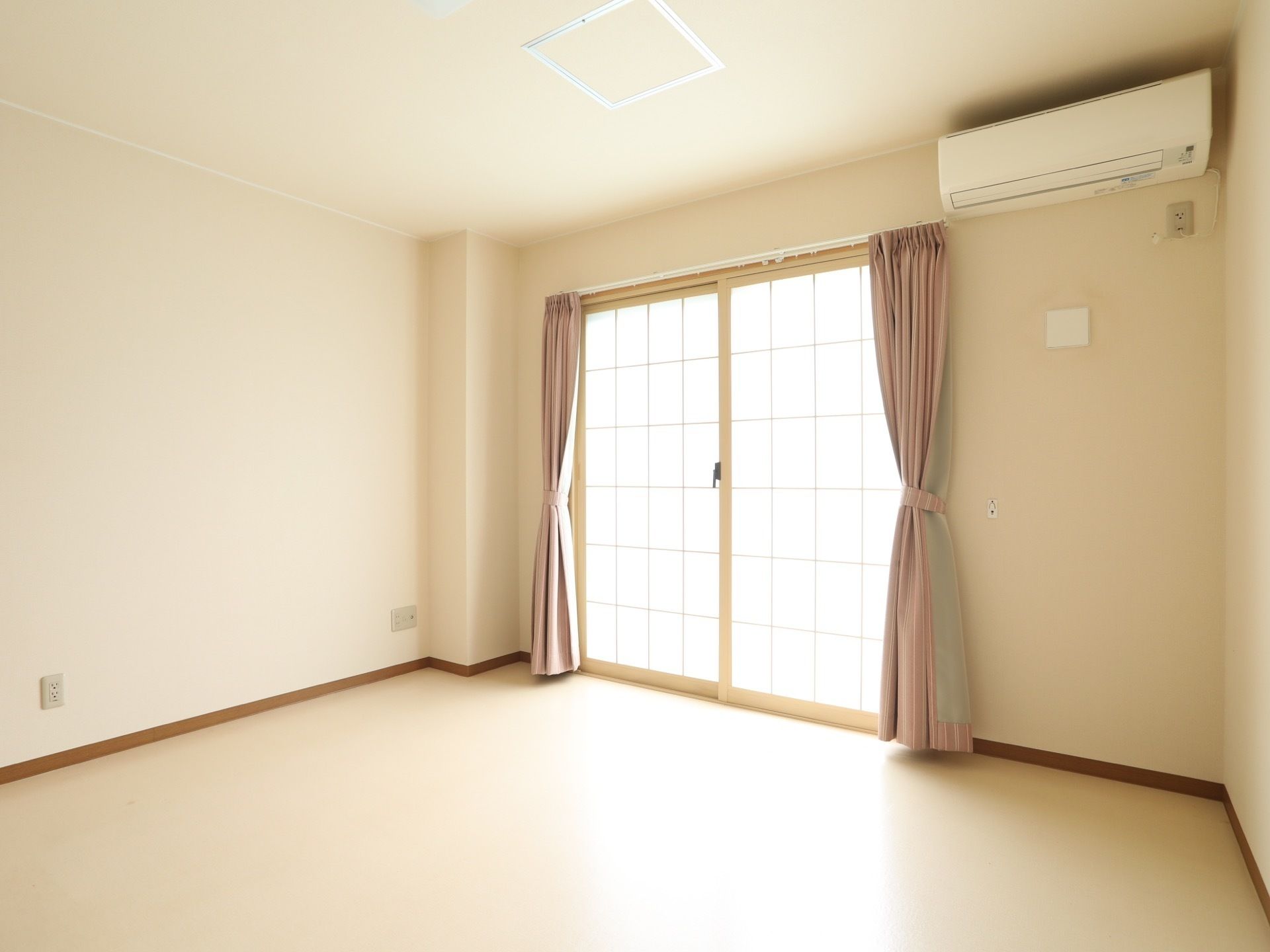 掃き出し窓から外光の入る居室。窓にはピンク色のカーテンが掛かっており、上にはエアコンが設置されている。