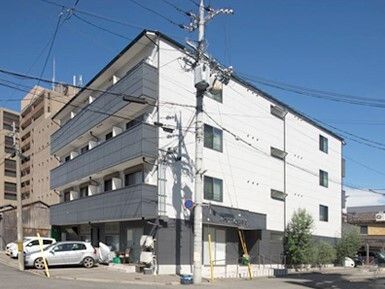 白い外壁の4階建ての建物の近くには複数の建物が建っていて、植物も植えられている。道路の面していて車も止められている。