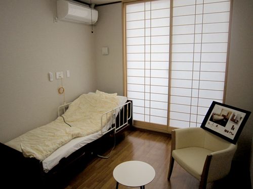 ベッドサイドには緊急通報装置が設置され、テーブルセットやエアコンが設置されたフローリングの部屋。