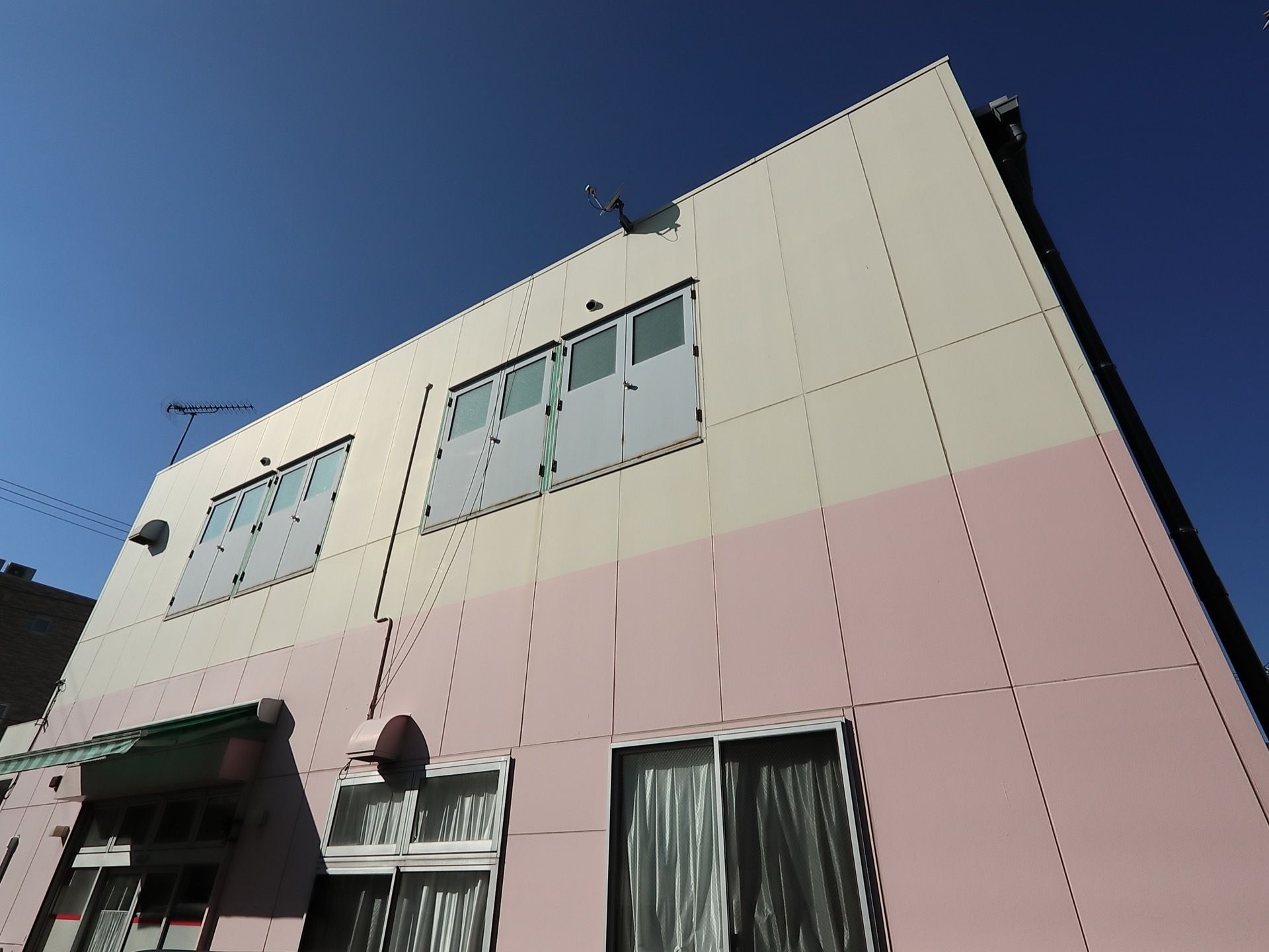 ピンク色と白の２色の壁の２階建ての建物である。カーテンの掛かった窓がある。玄関部分にはルーフがついている。