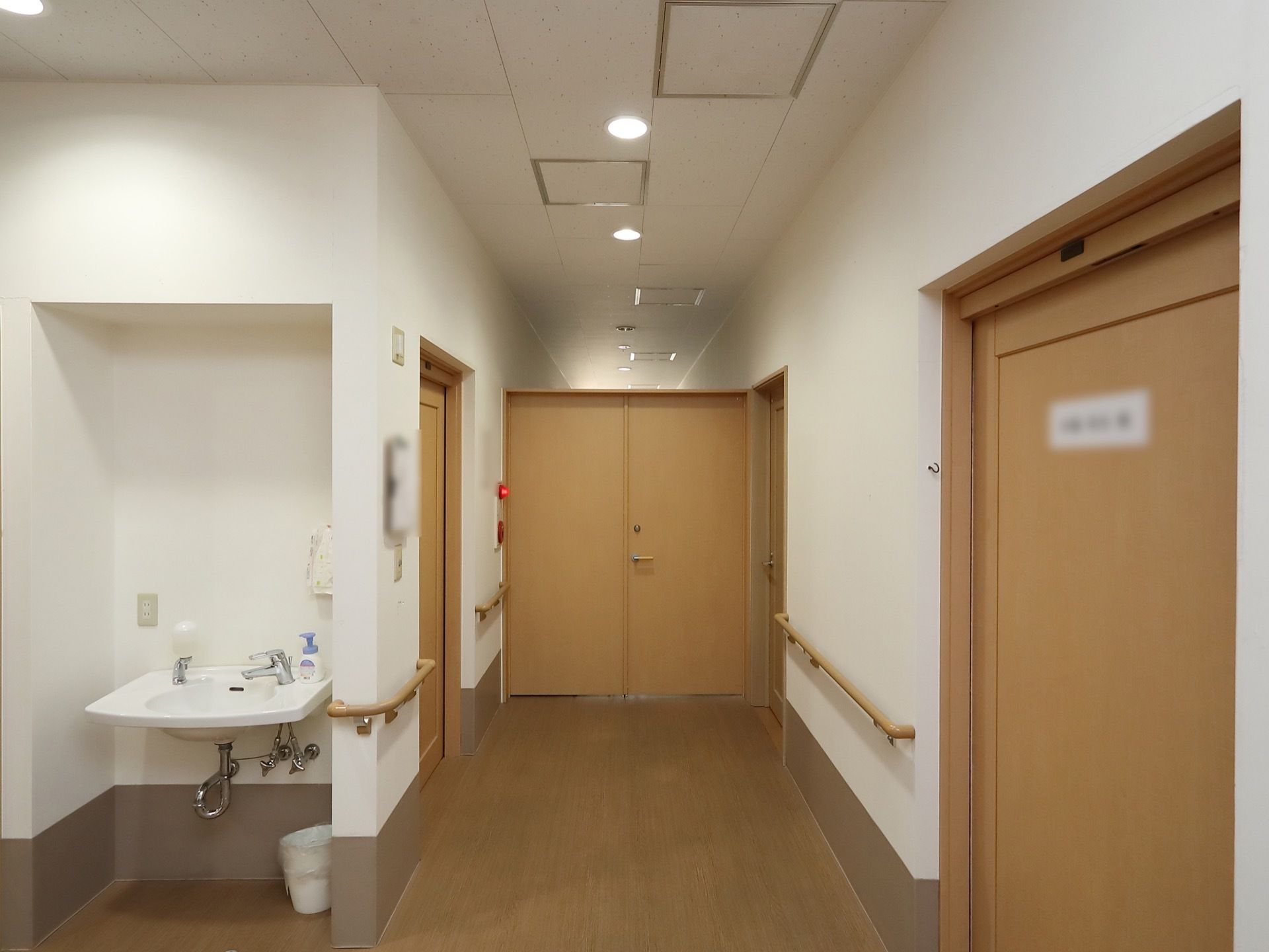 木目調の床とドアが並んでいる廊下がある。壁には手すりが設置されており、側の奥まった場所には洗面台がある。