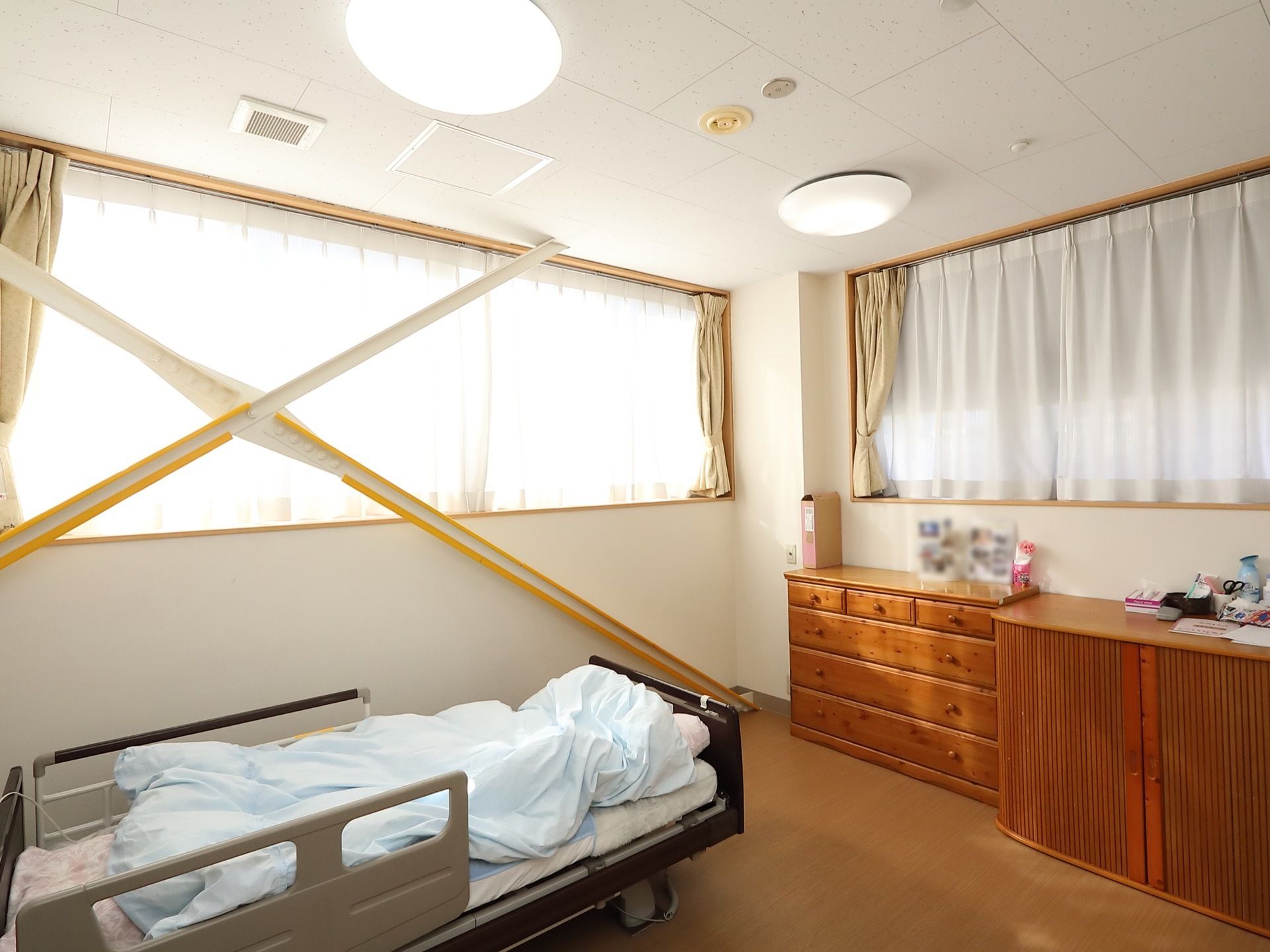大きな窓が２つついている角部屋の居室がある。タンスがあり、上には物が置かれている。介護用ベッドがある。
