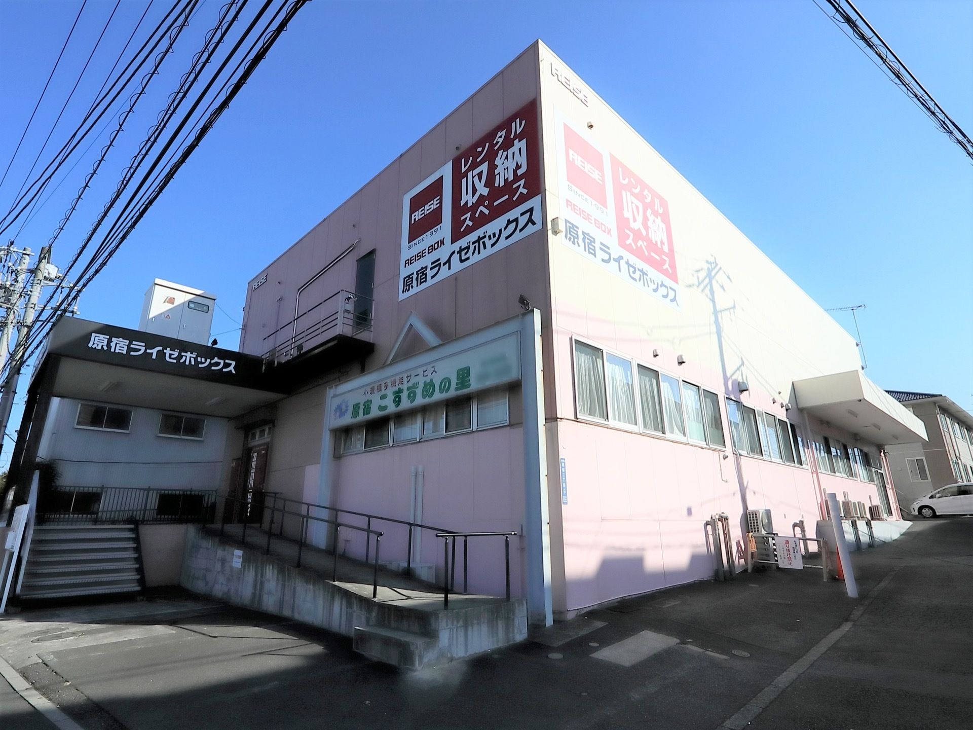 外観がピンクと白で、２階建ての大きな建物である。２階部分には他店舗の看板が掛かっている。玄関までスロープと手すりが設置されている。