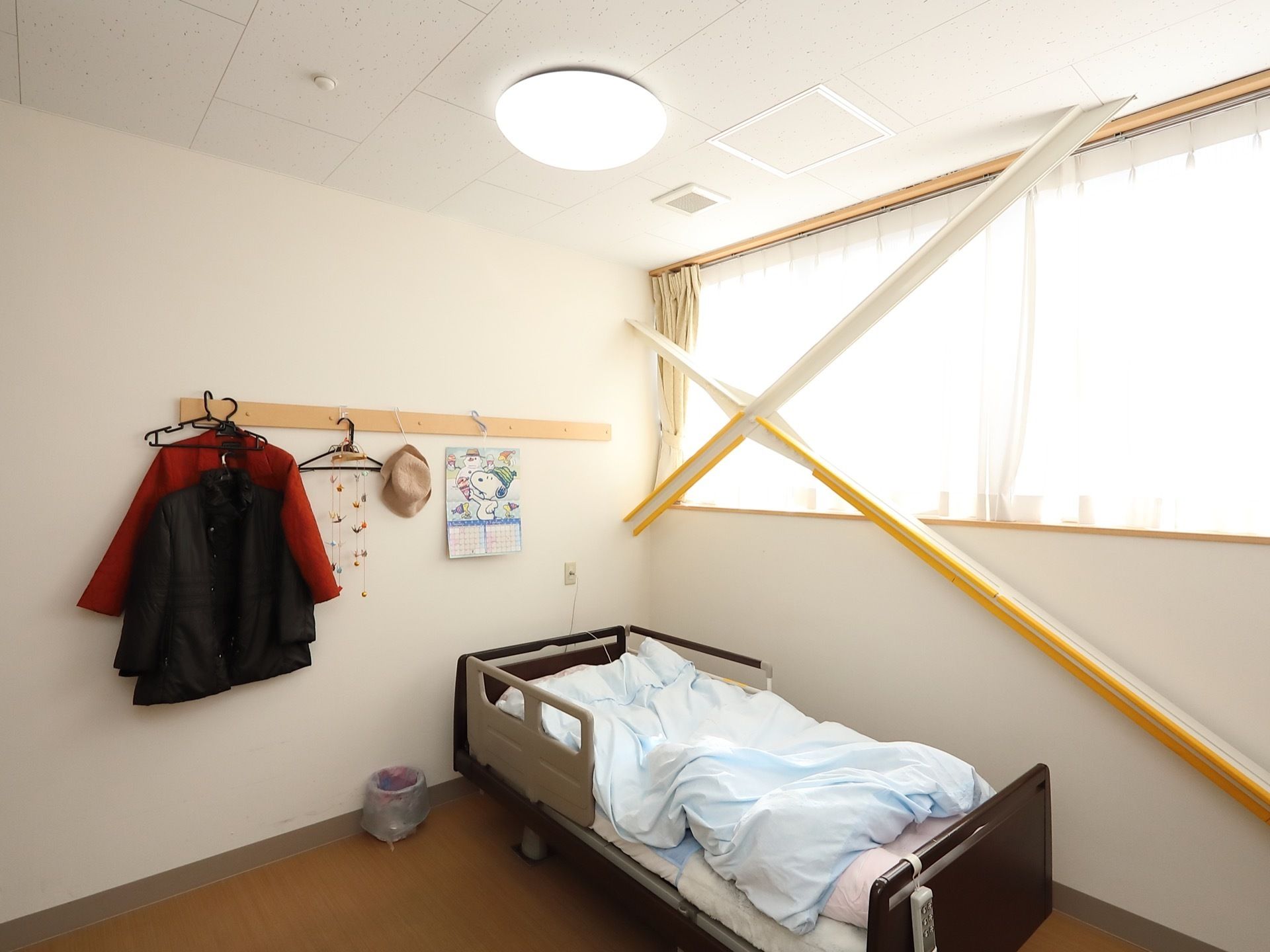 壁の上半分が大きな窓になっている明るい居室がある。介護用ベッドが置かれ、壁には物を掛けるスペースがある。