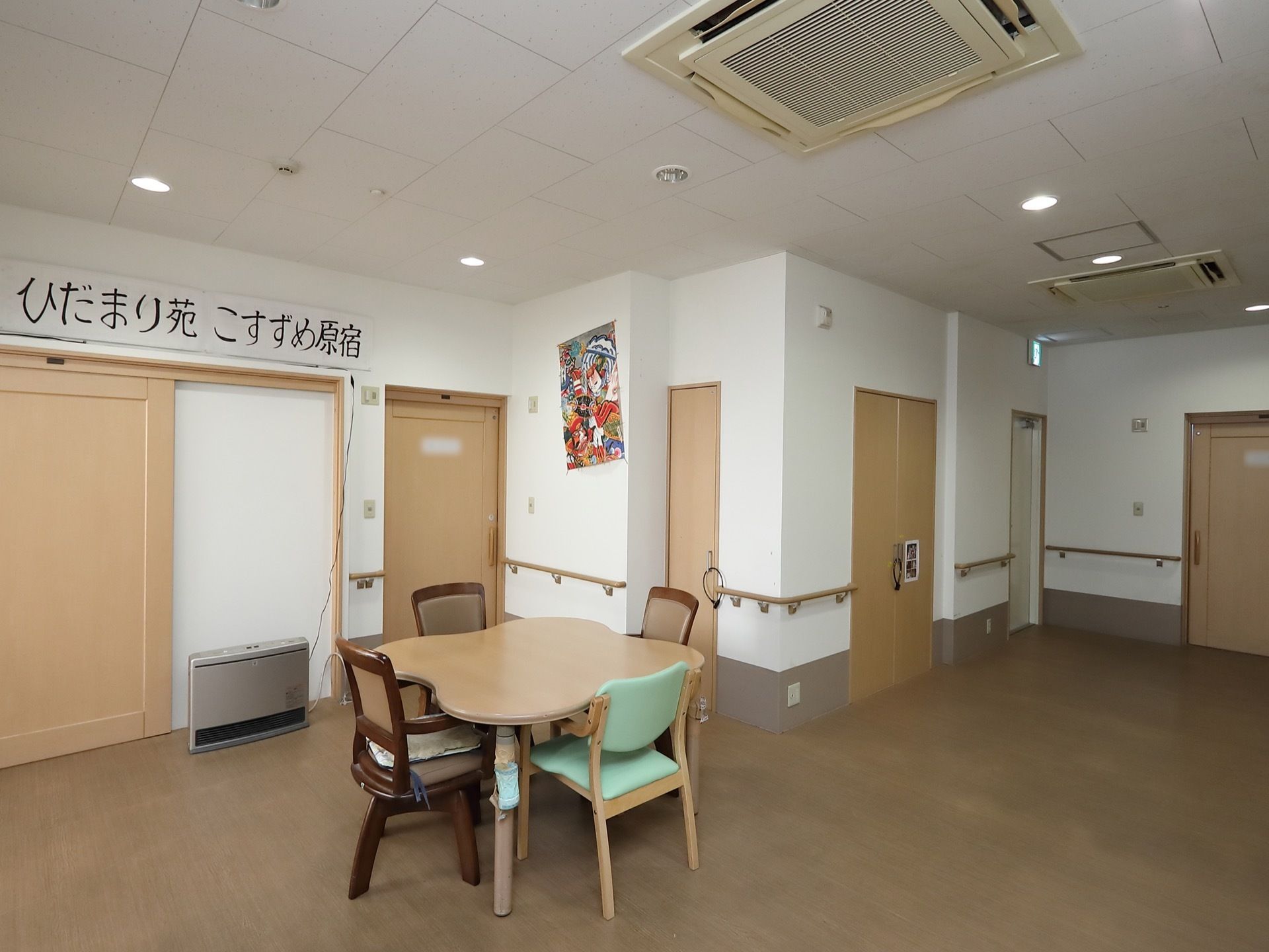 居室のドアが並んでいる広いスペースにある談話室。テーブルと椅子が置かれ、壁にはポスターと施設名が書かれた看板が掛けられている。