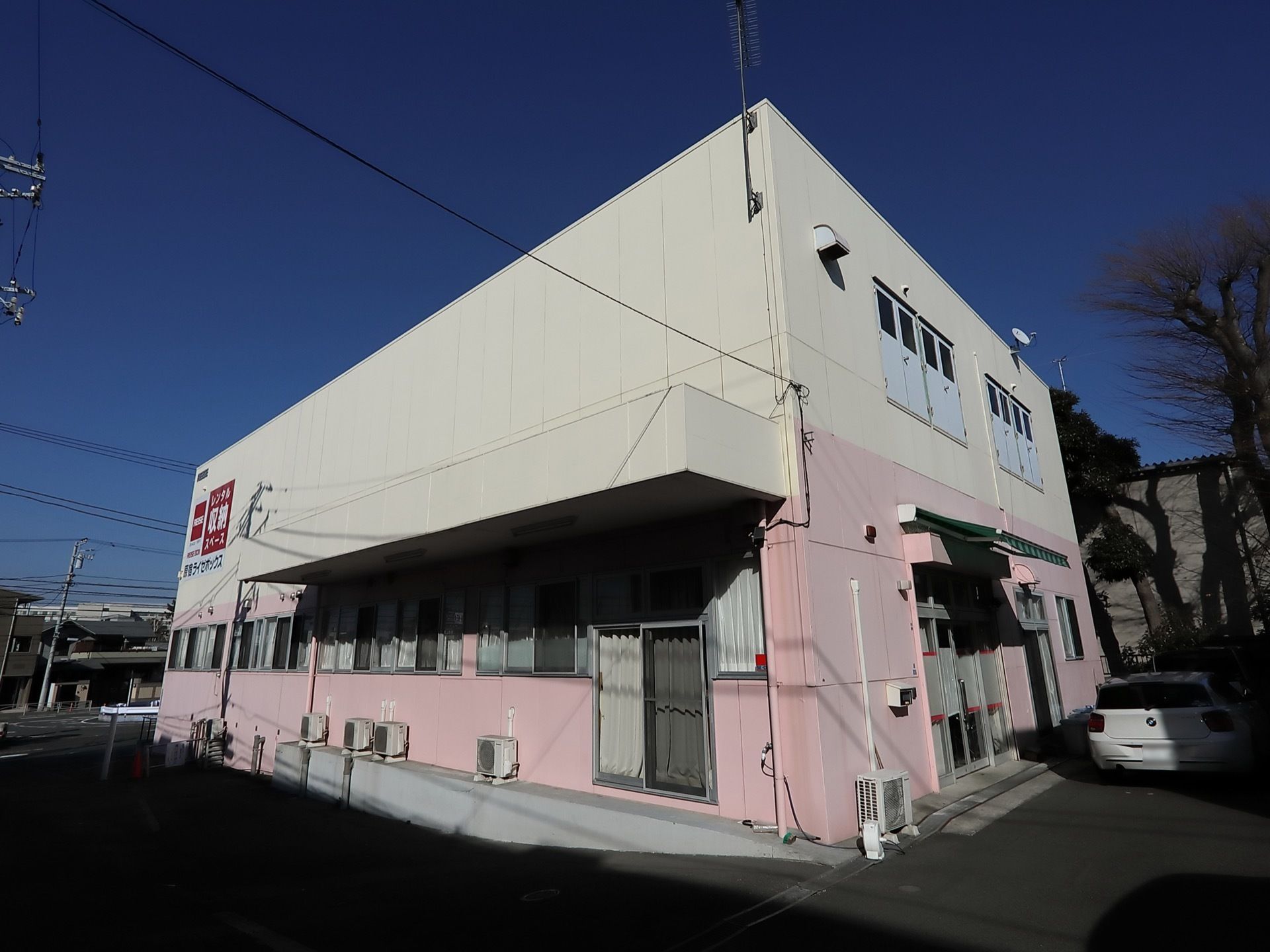 １階部分が白、２階部分がピンクに塗られた壁の四角い建物がある。窓にはカーテンが掛けられており、日当たりが良い立地にある。