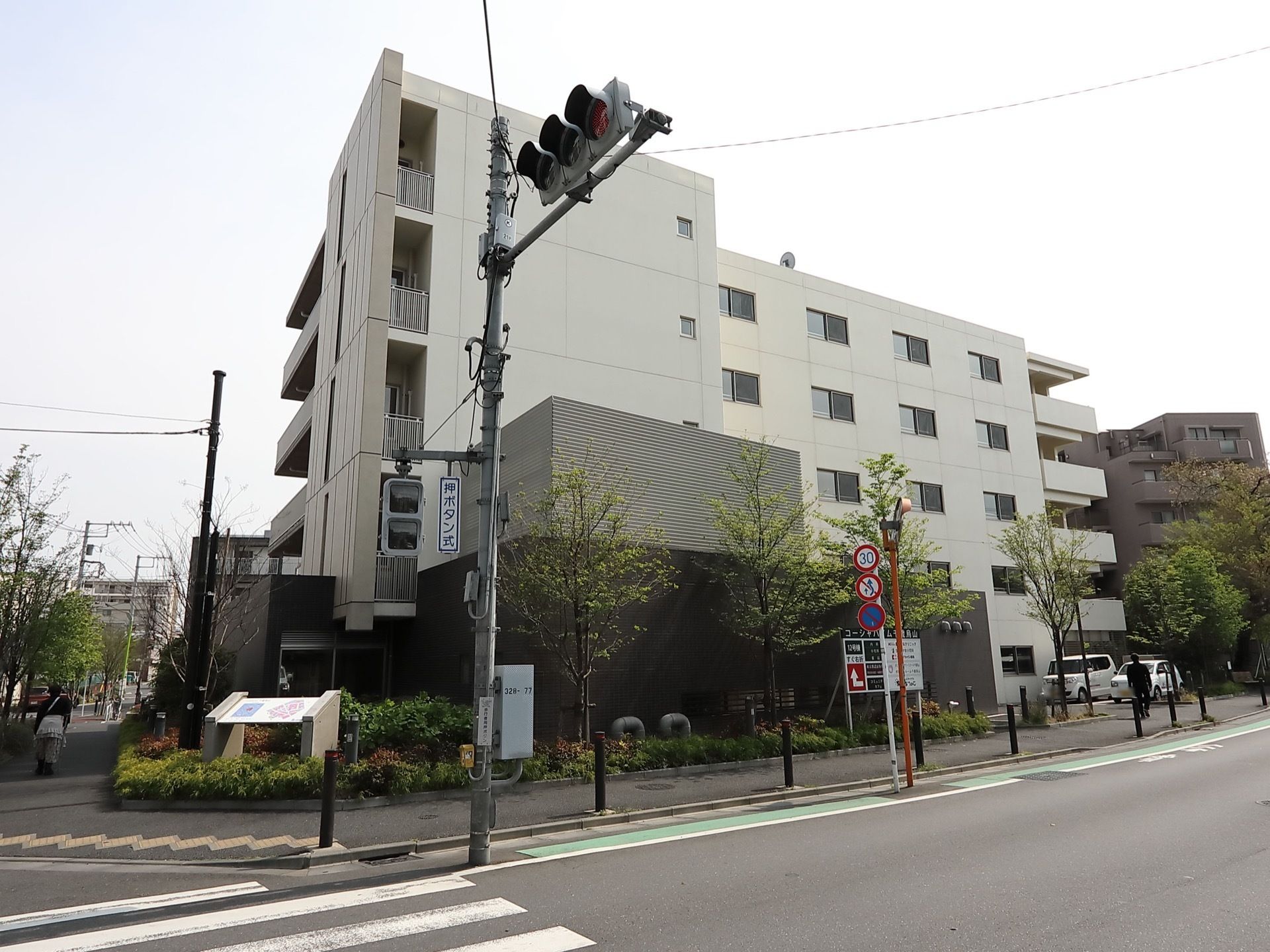 交差点の角地に立っており、施設前には信号機がある。白い外壁で５階建ての建物は周囲を樹木に囲まれている。