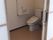 手すりや呼び出しボタンが付いたトイレは広く、シンプルな便座が付き、スライド式のドアが設置されている。