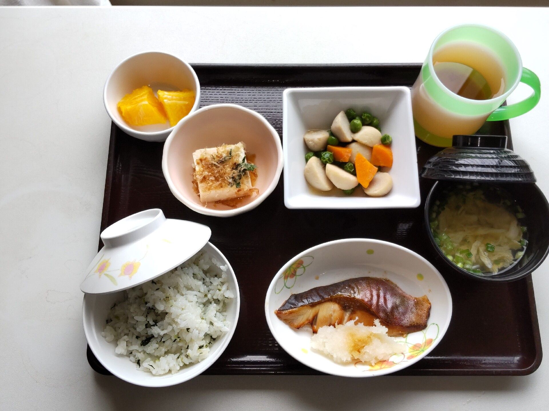 野菜の煮物や冷ややっこ、わかめご飯に魚の煮つけ、みそ汁などが並べられた黒いトレー。