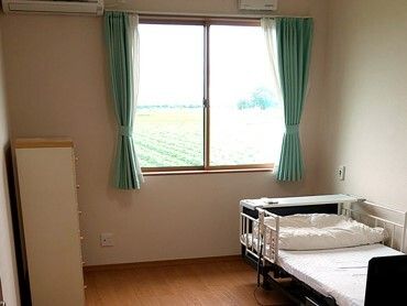 レースカーテンとカーテンが付いた大きな窓があるフローリングの部屋には寝具付きのベッドが置かれ、タンスもある。