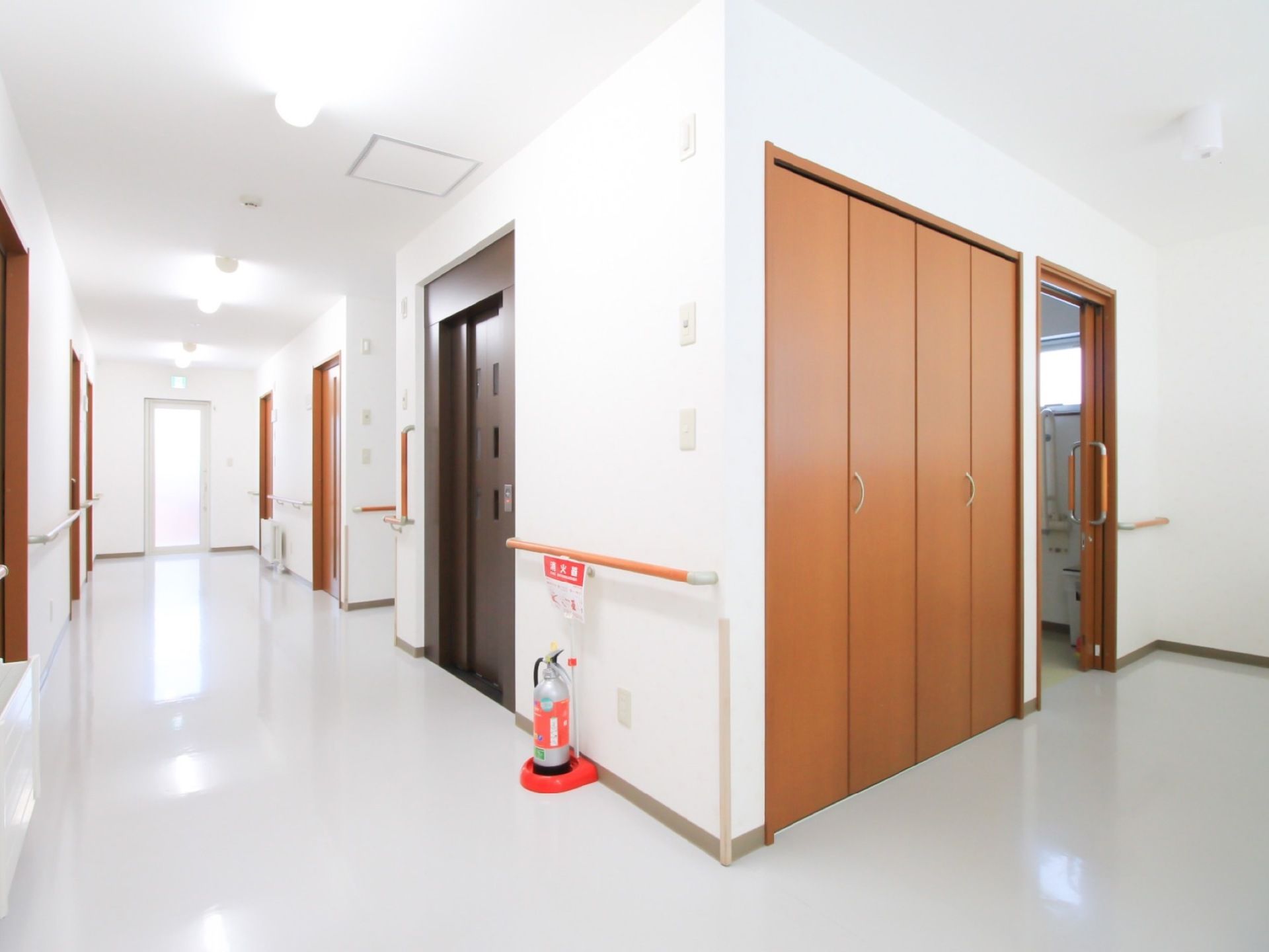 白色を基調としたすっきりとした廊下である。エレベーター乗降口のすぐそばの手すりの下に消火器が置かれている。