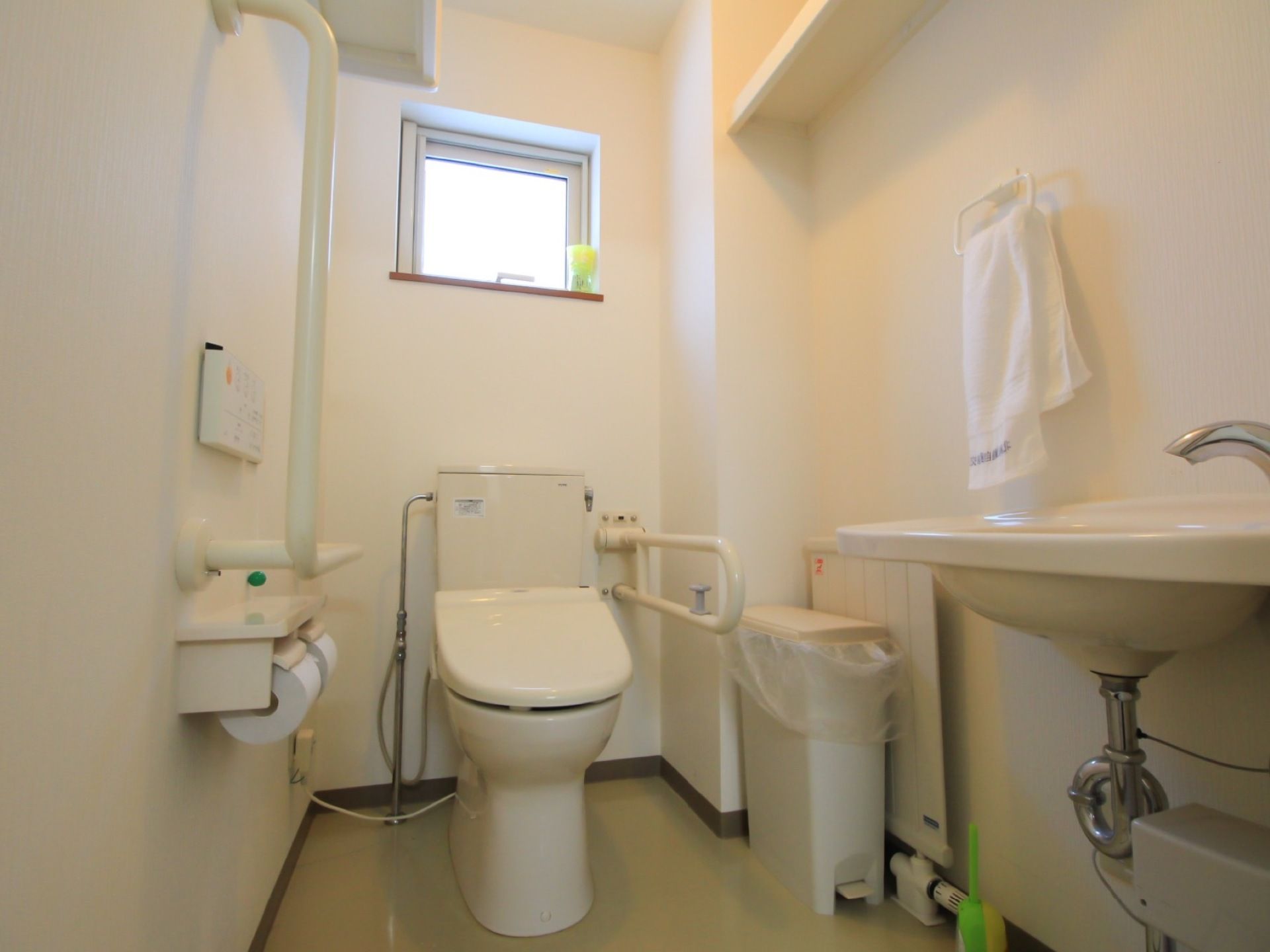 白色で統一されたトイレである。壁に大きなL字型の手すりが設置され、その近くに便座機能の操作パネルがある。