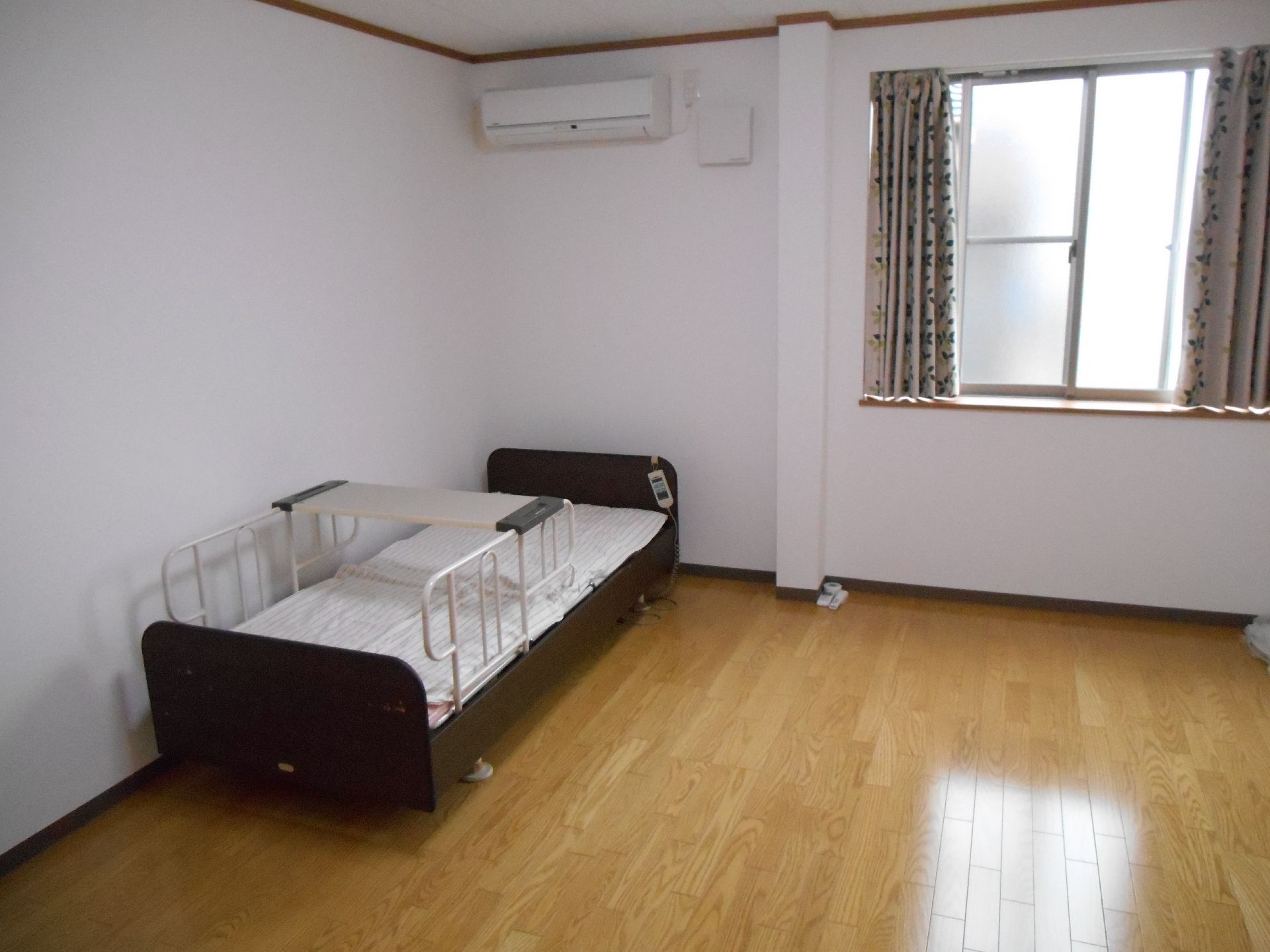 カーテンが付いた窓があるフローリングの部屋には寝具付きのベッドやエアコンがあり、家具はほかにはない。