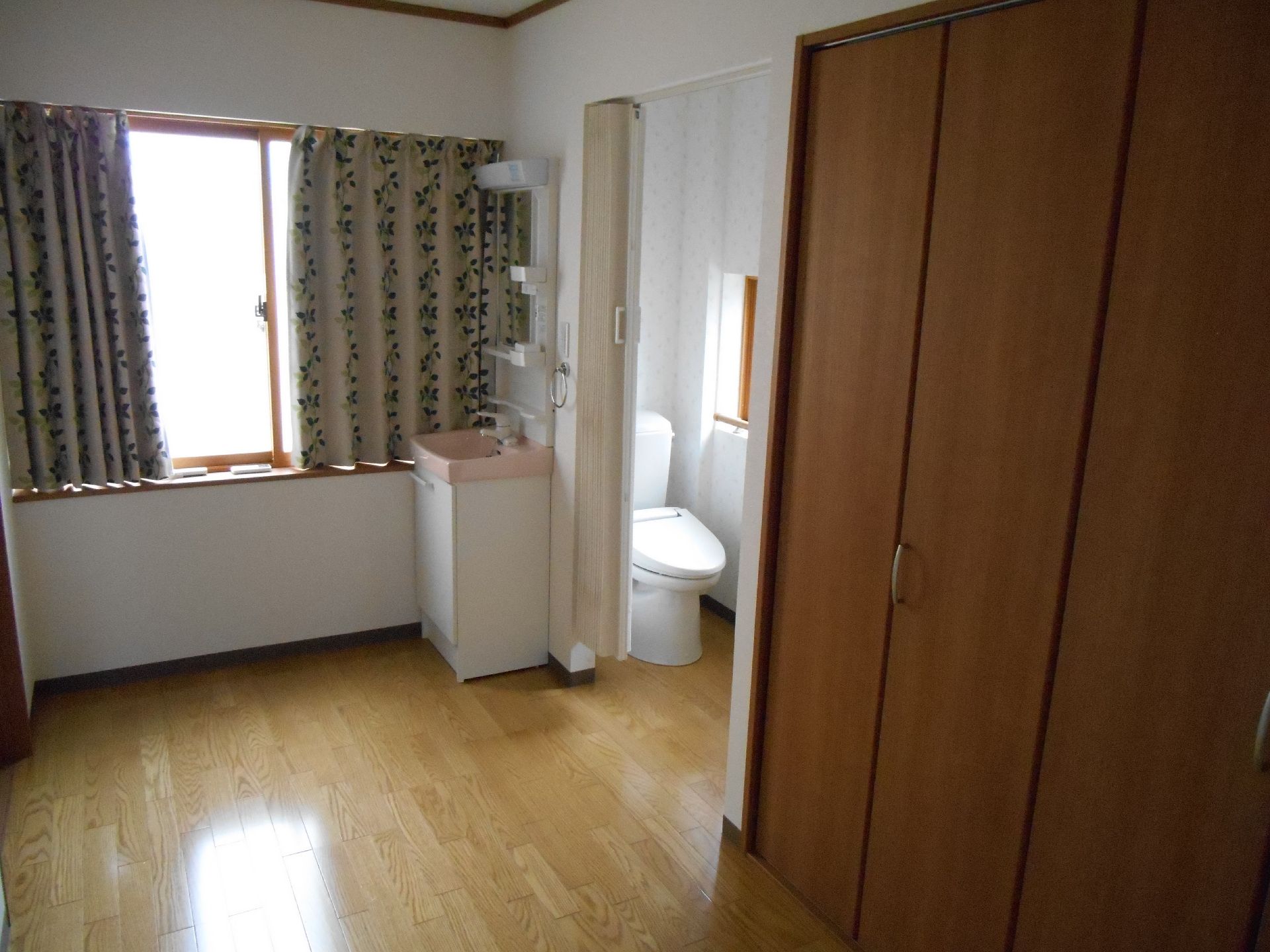 カーテンで開け閉めするタイプのトイレが付いたフローリングの部屋には、大きなクローゼットがあり、窓にはカーテンが付いている。