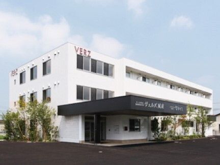 3階建ての建物は白を基調としていて、赤い文字で住宅名が書かれている。黒い屋根が玄関に付き、周りには緑も植えられている。