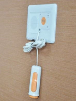 オレンジと白の緊急通報装置が木目調の壁についている。オレンジのボタンを押せば通話ができるタイプの装置で長いコードも付いている。