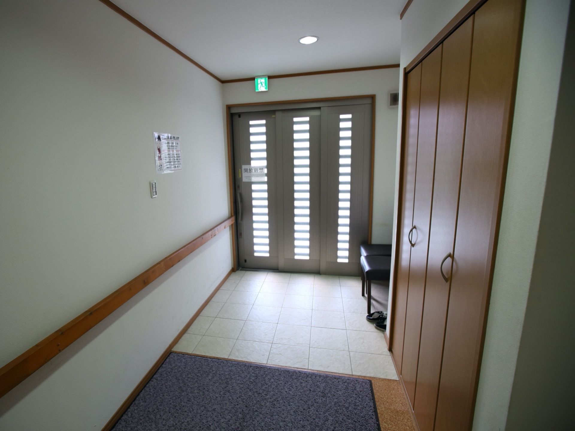 スライド式の扉の玄関で、ドアノブの近くに白い張り紙が貼られている。右側に黒い椅子が２脚並べて置かれている。