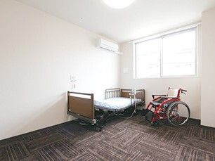じゅうたんが敷かれたワンルームタイプの部屋。ベッドと車いすが置いてある様子