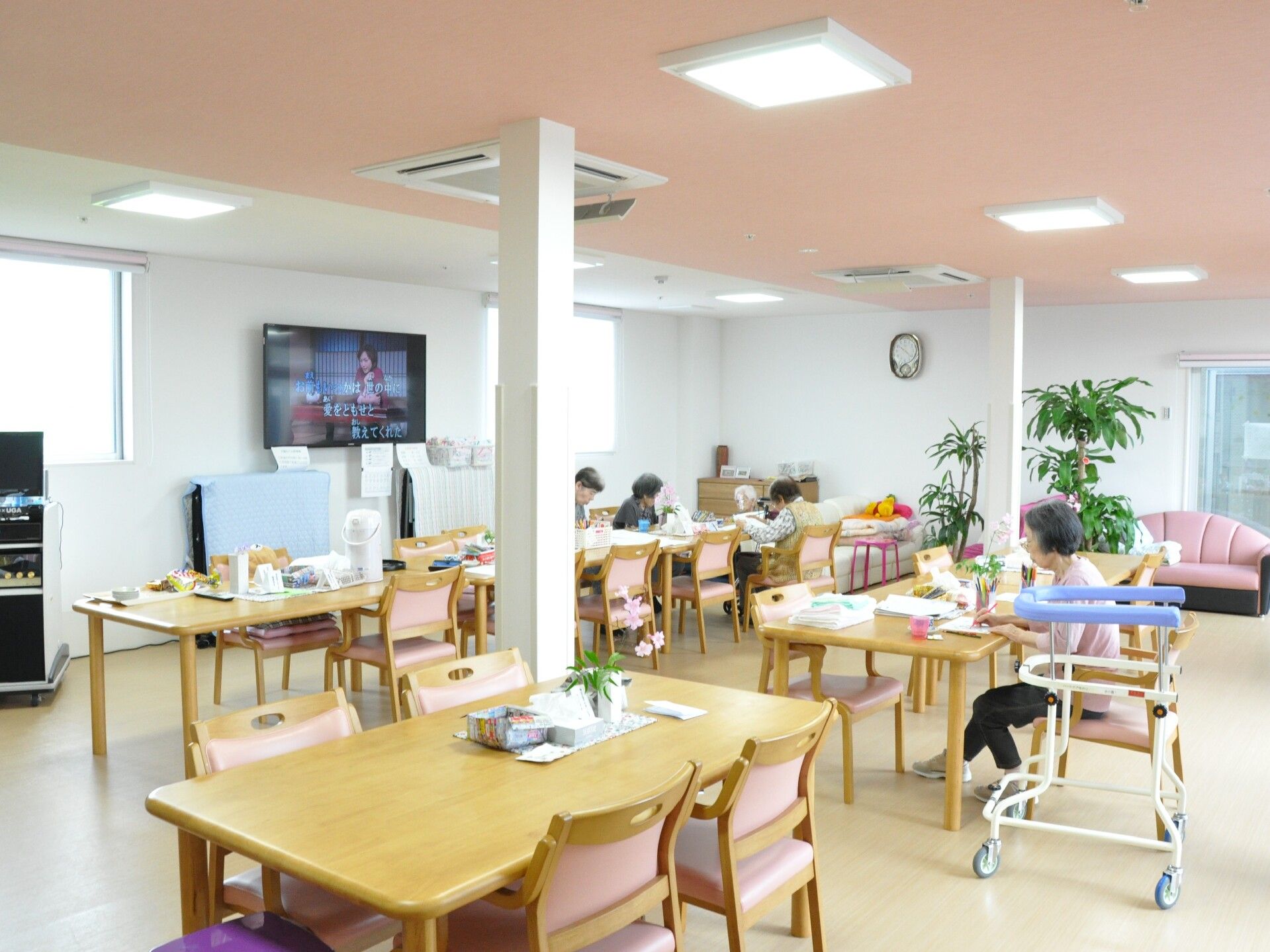 4人掛けのダイニングテーブルが複数置かれた食堂。歩行器が置かれ、数名の入居者が思い思いの場所で過ごしている。